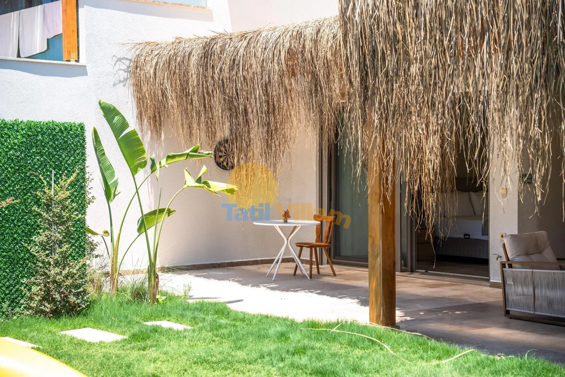 Un patio con techo de paja junto a un pequeño césped tiene una mesa y asientos.