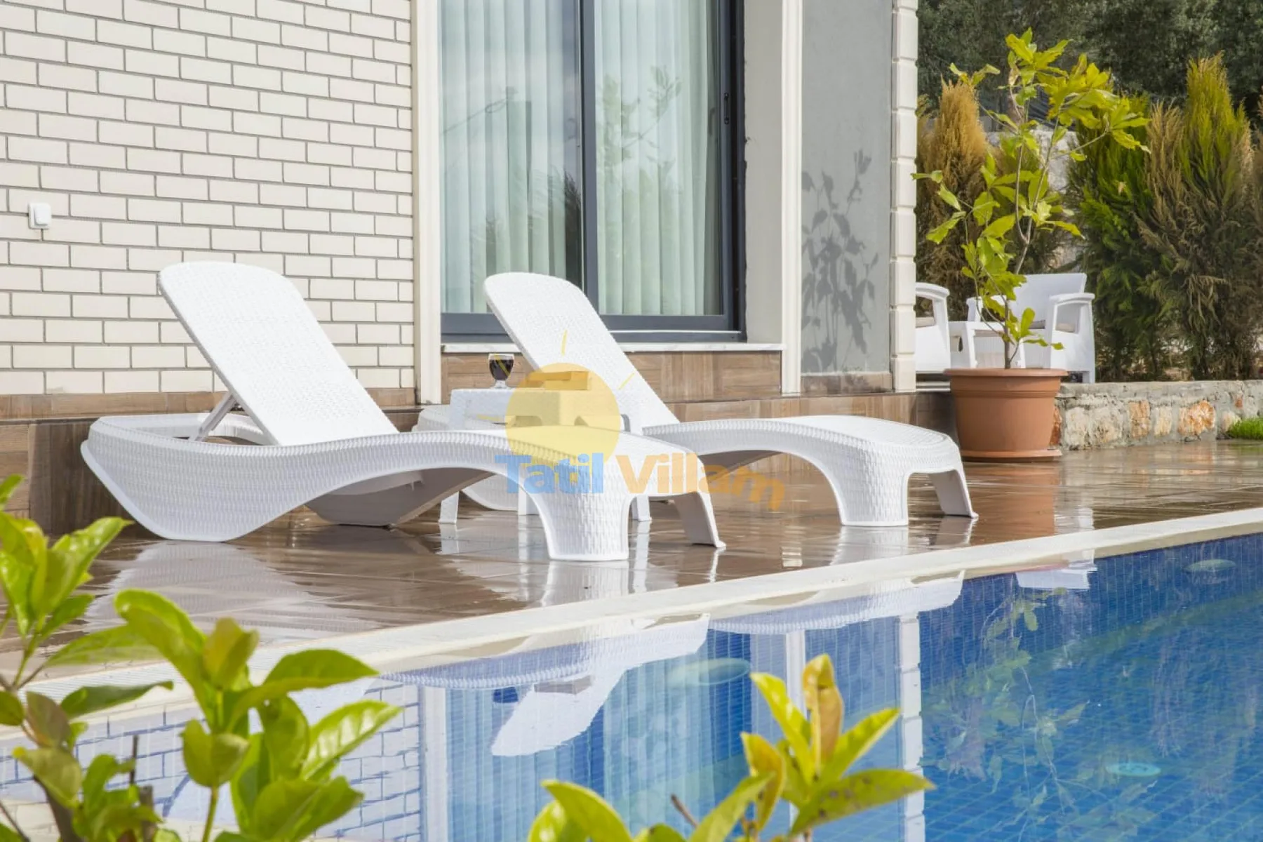 Poolside seating sits beside a blue swimming pool and a light brick wall.