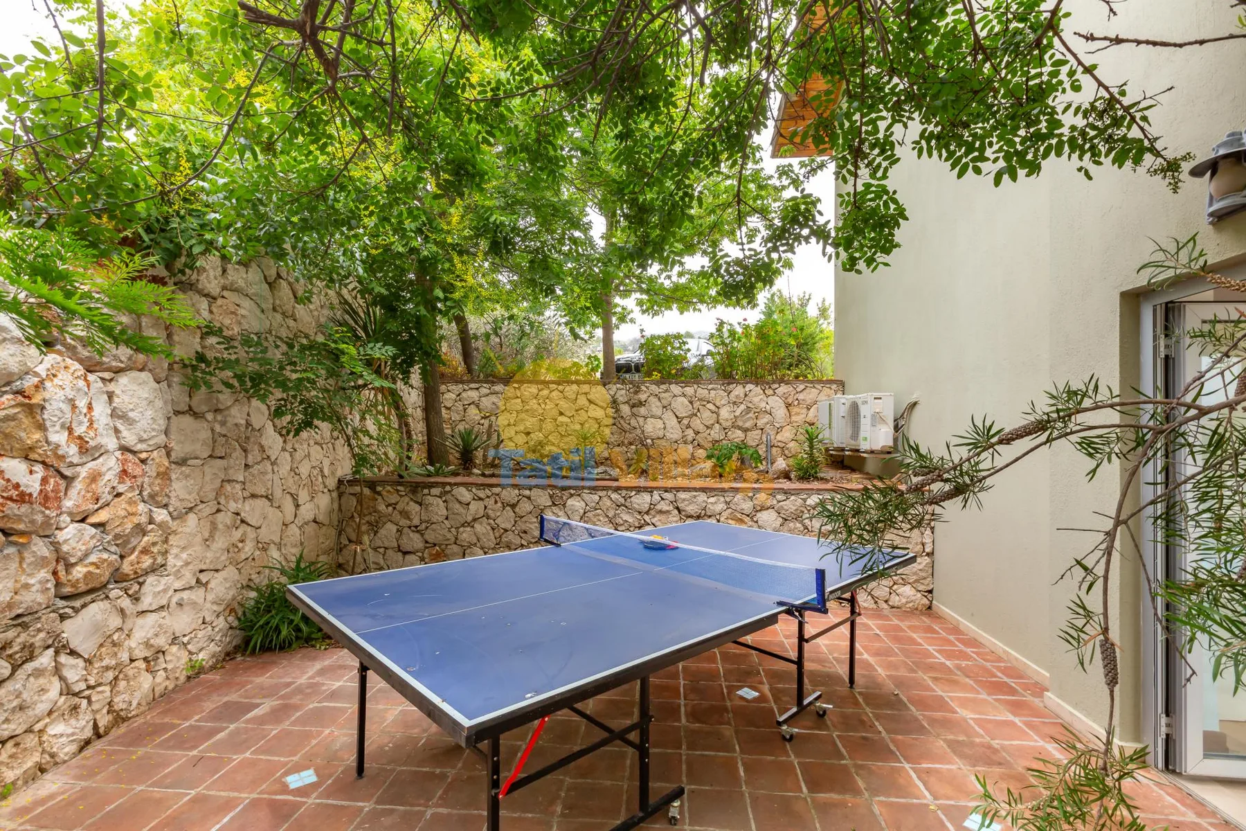 An outdoor table tennis area sits on a tiled patio beneath leafy trees.