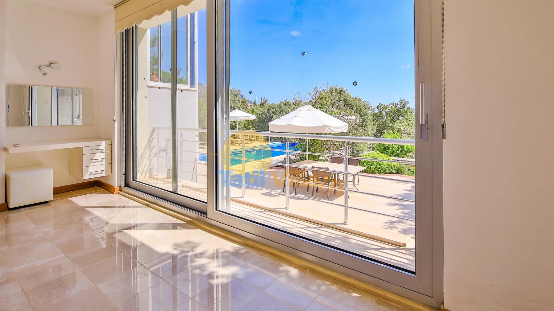 A bright room opens onto a balcony with outdoor seating and a pool view.