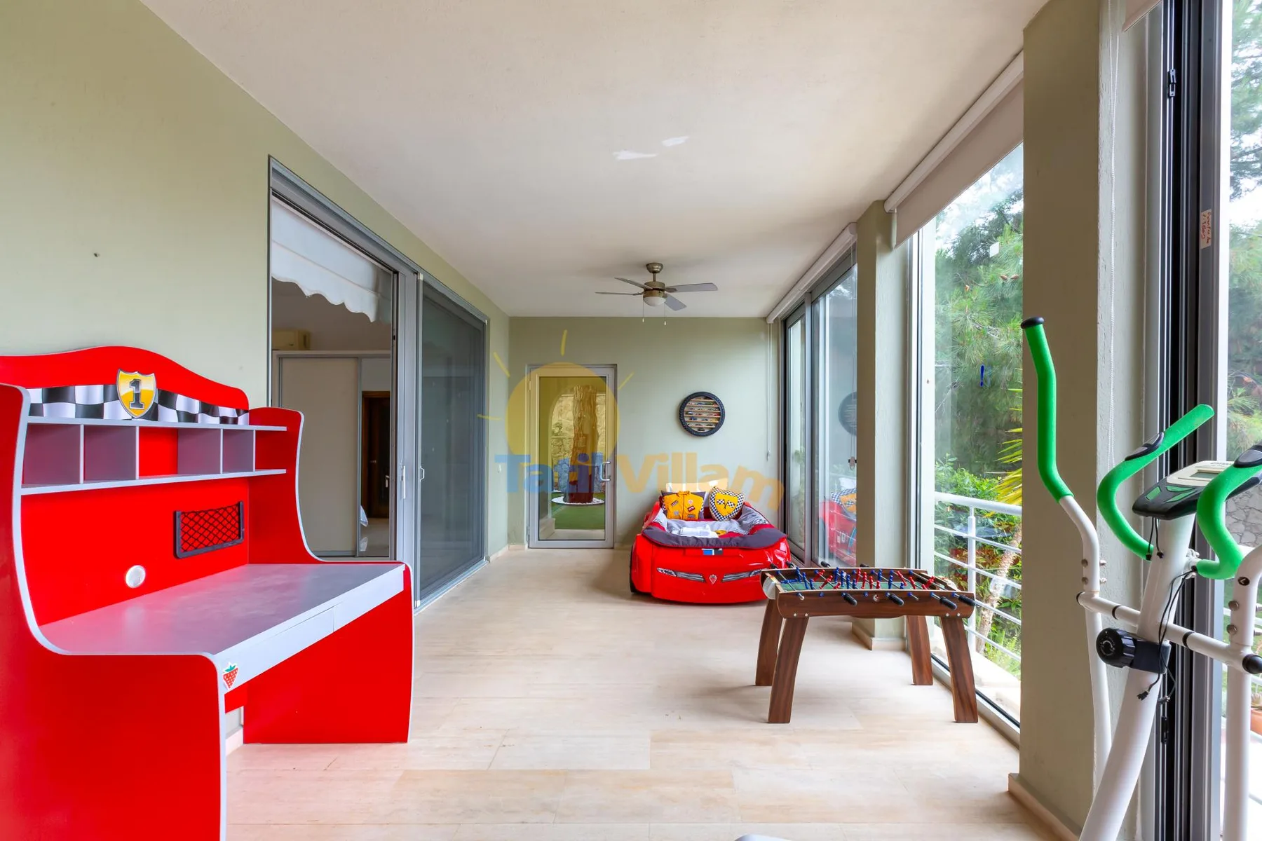 A bright enclosed patio has large windows, a foosball table, and exercise equipment.