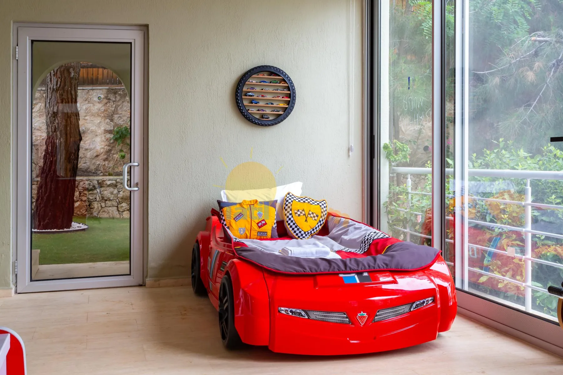 A child’s bedroom features a red car-shaped bed beside large windows.