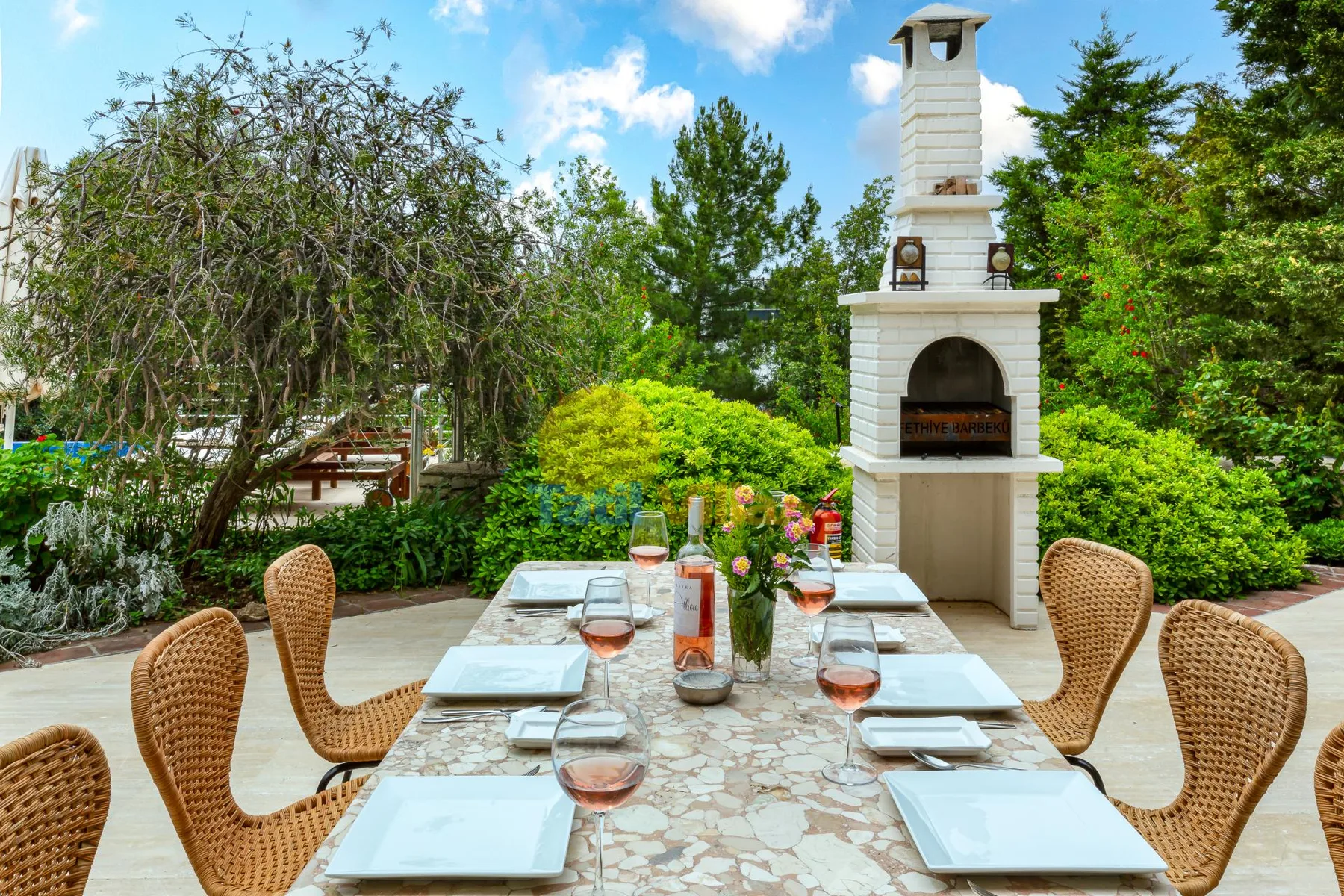 An outdoor dining table is set beside a stone barbecue in a leafy garden.