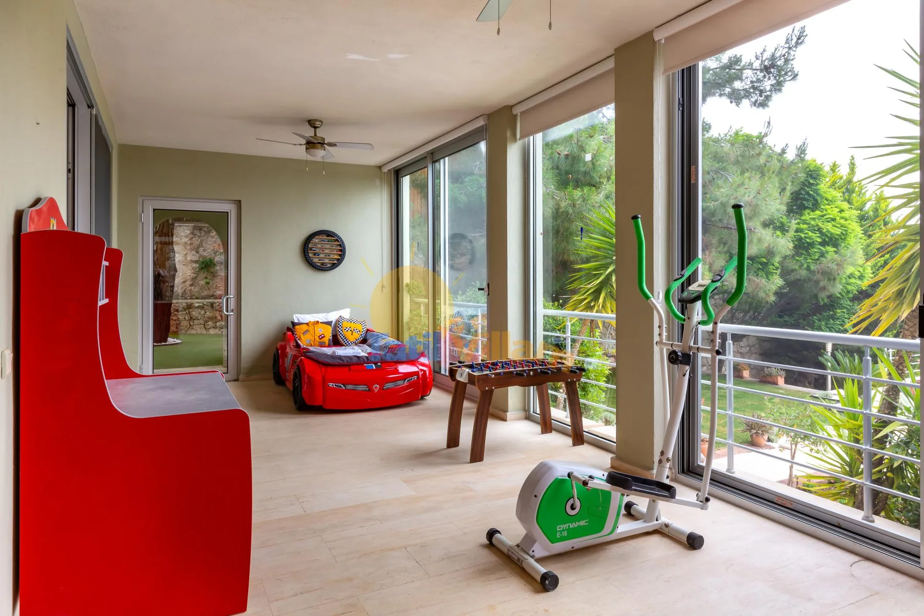 A bright enclosed patio has large windows, exercise equipment, and a small game table.