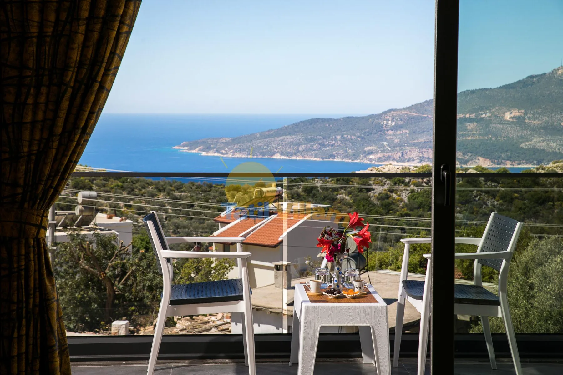 A balcony seating area overlooks the sea and a coastal hillside.