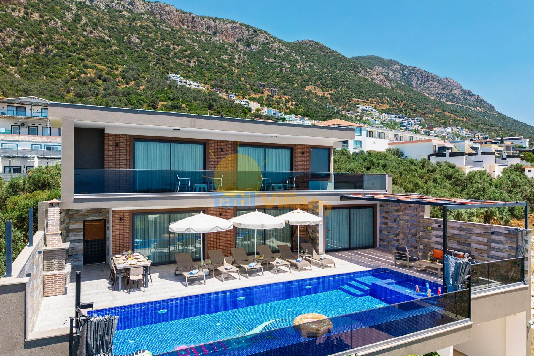 A modern hillside villa overlooks a swimming pool and outdoor seating area.