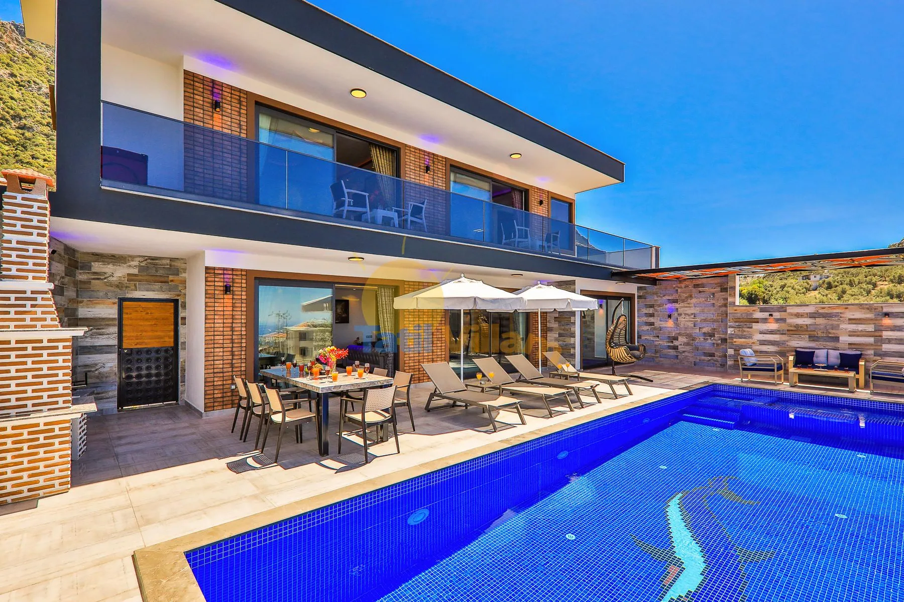 A modern house opens onto a swimming pool, with outdoor seating beneath umbrellas.