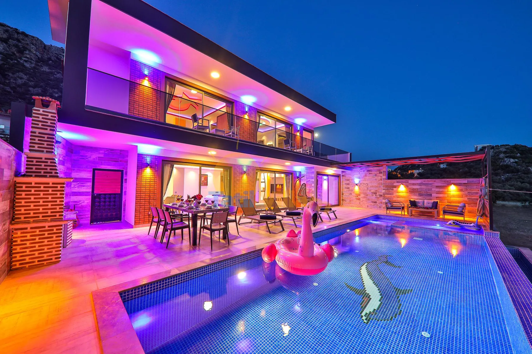 A modern house opens onto a swimming pool and outdoor dining area at night.