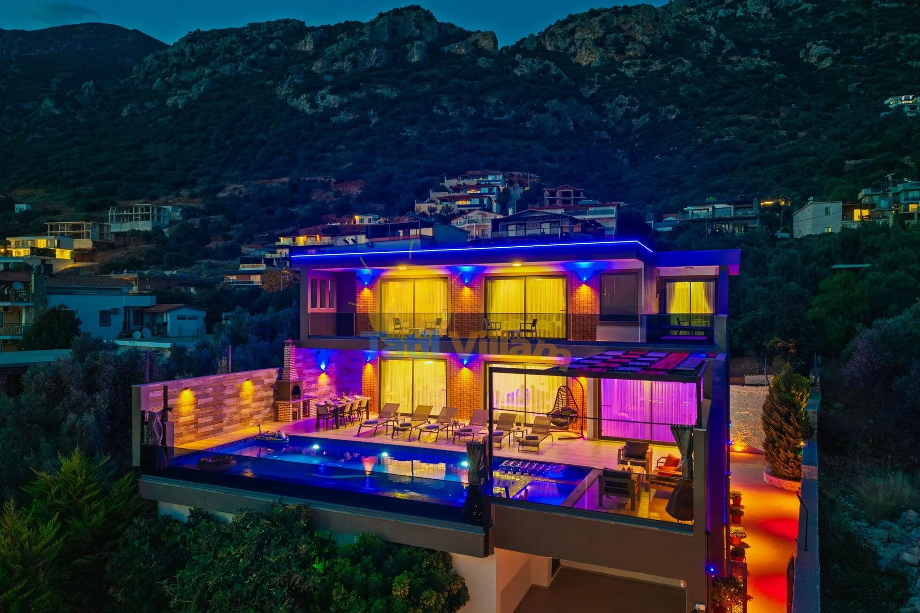 A brightly lit modern house with a swimming pool sits below a dark mountain ridge.
