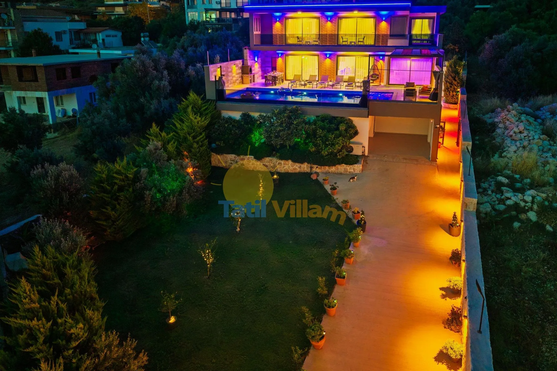 A brightly illuminated multi-story house stands beside a landscaped garden and driveway.