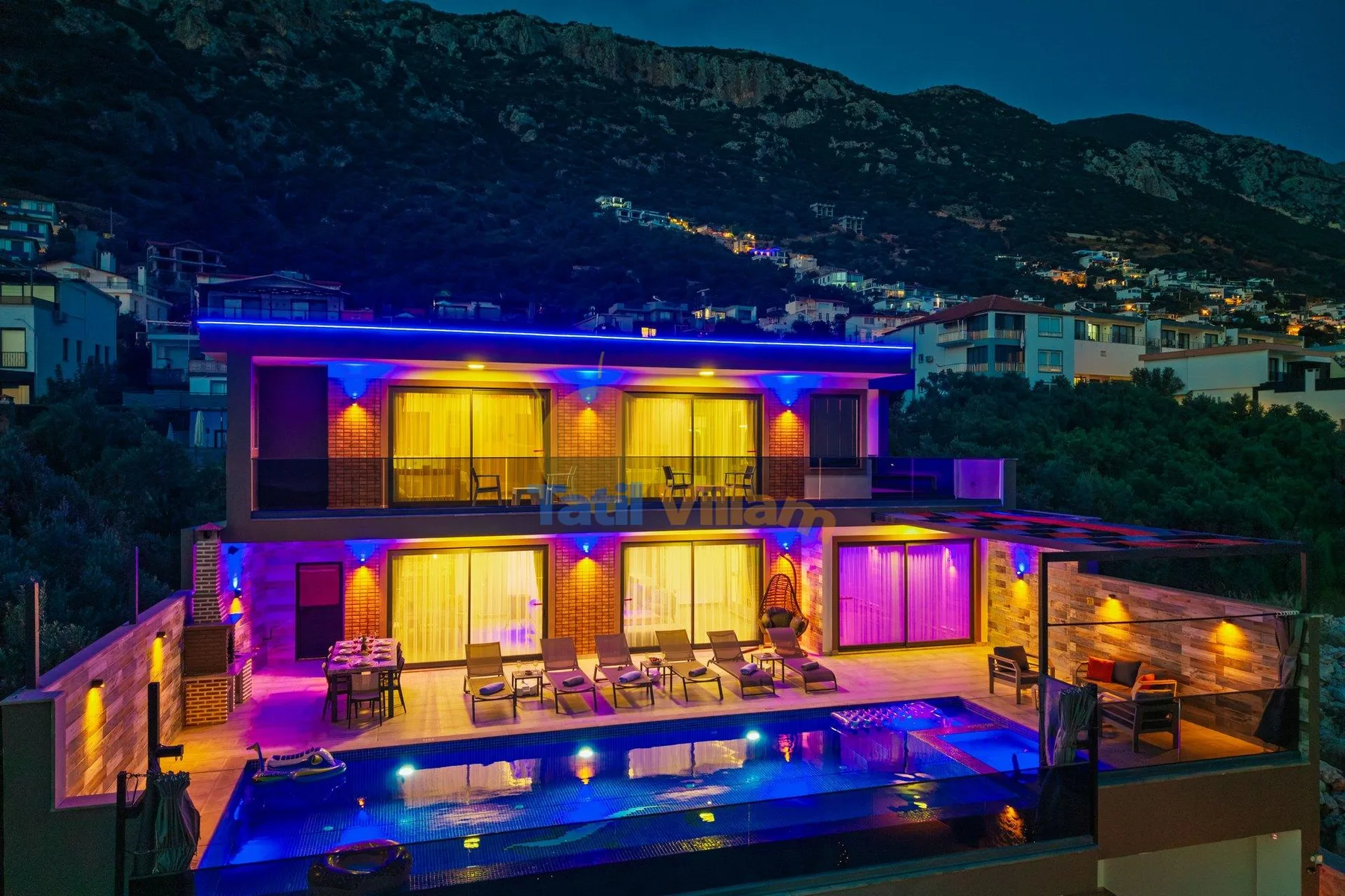 A modern house glows with colorful lights beside a swimming pool at night.