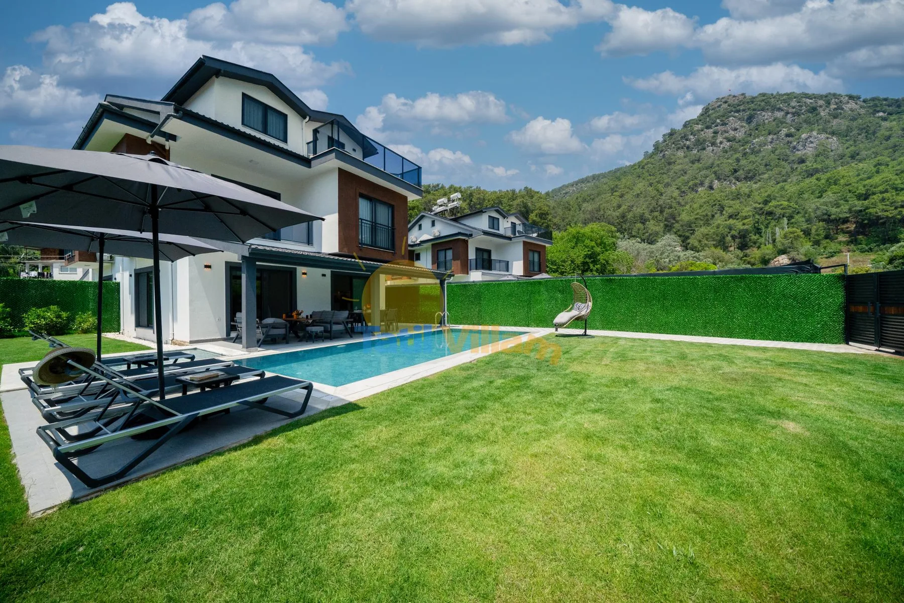 Una casa moderna da a un césped y una piscina, con colinas al fondo.