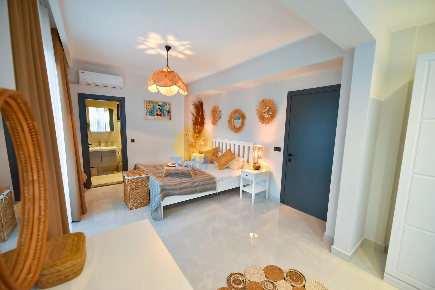 A bright bedroom has a white bed with patterned bedding, woven wall decorations, and a dark door.