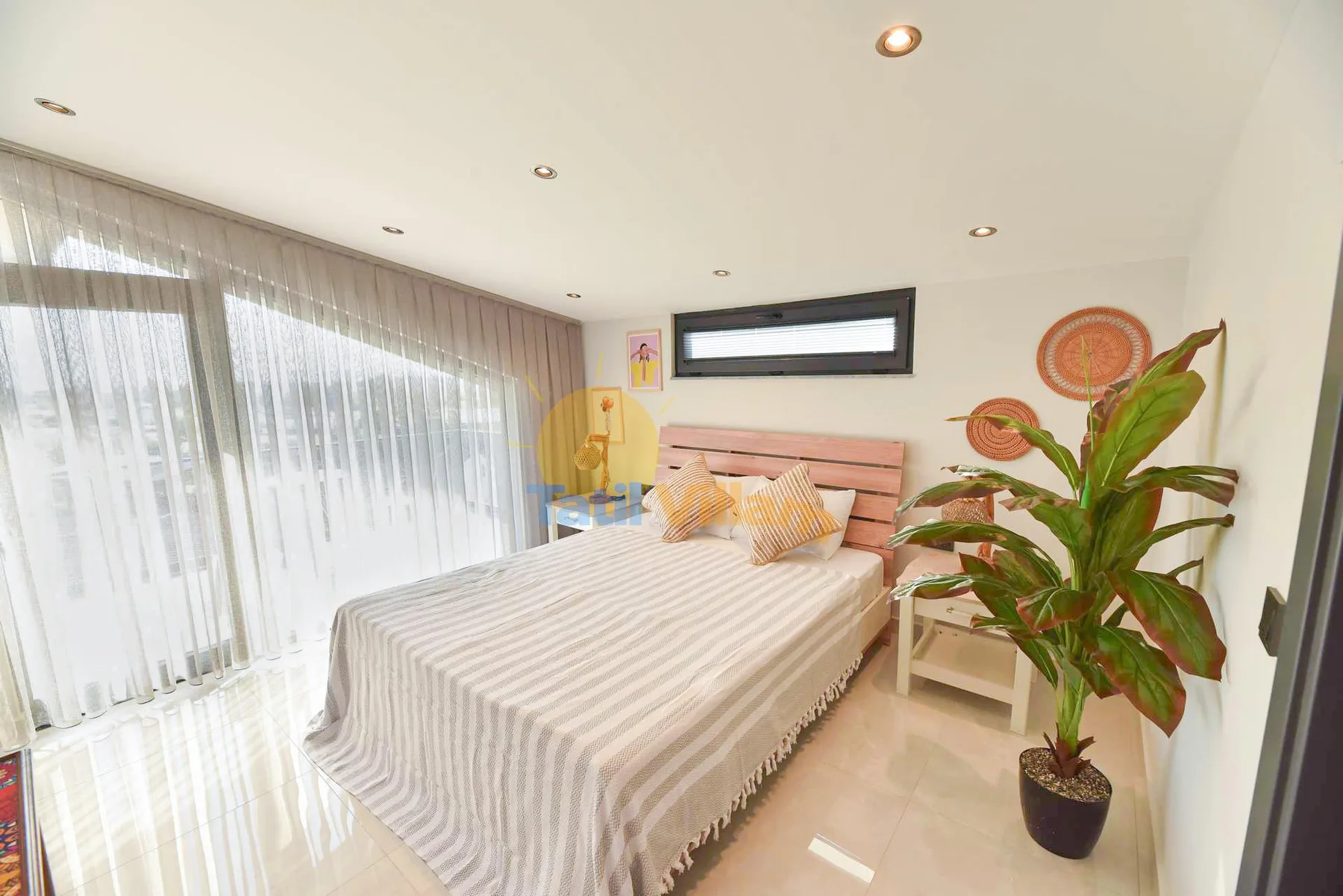 A bright bedroom has a striped double bed, sheer curtains, and a leafy potted plant beside the bed.