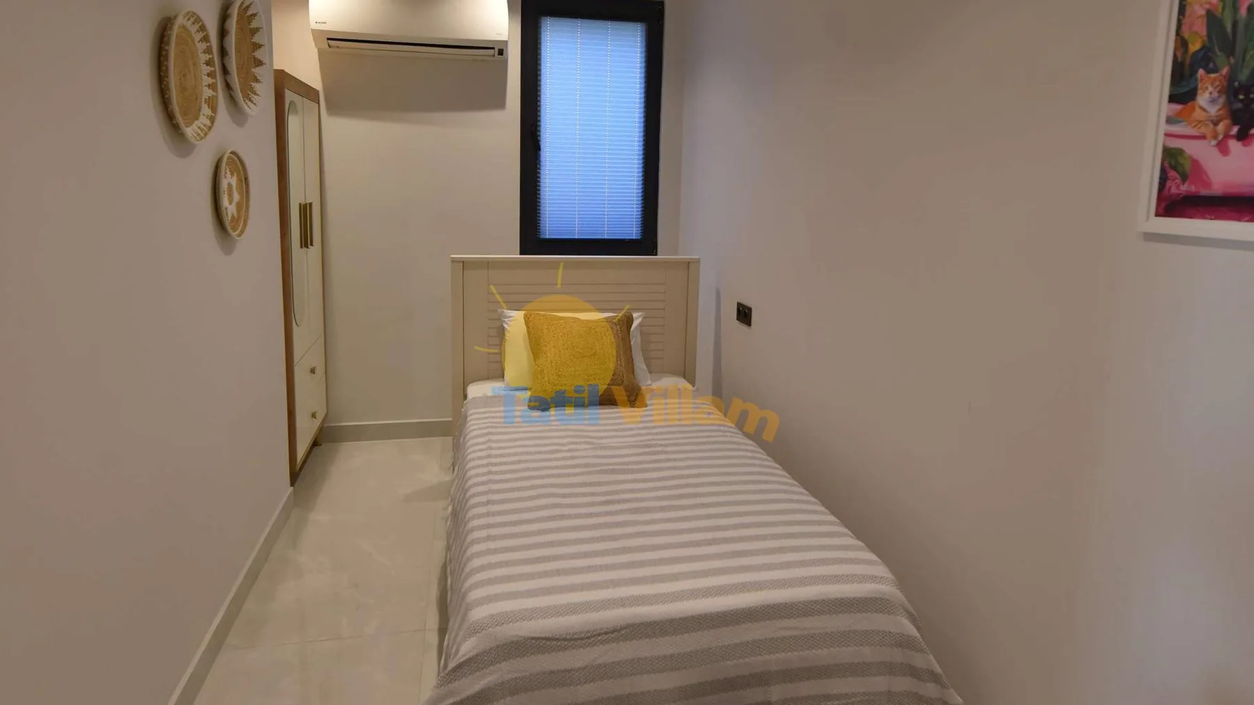 A narrow bedroom contains a single bed with striped bedding, a window, and a wall-mounted air conditioner.