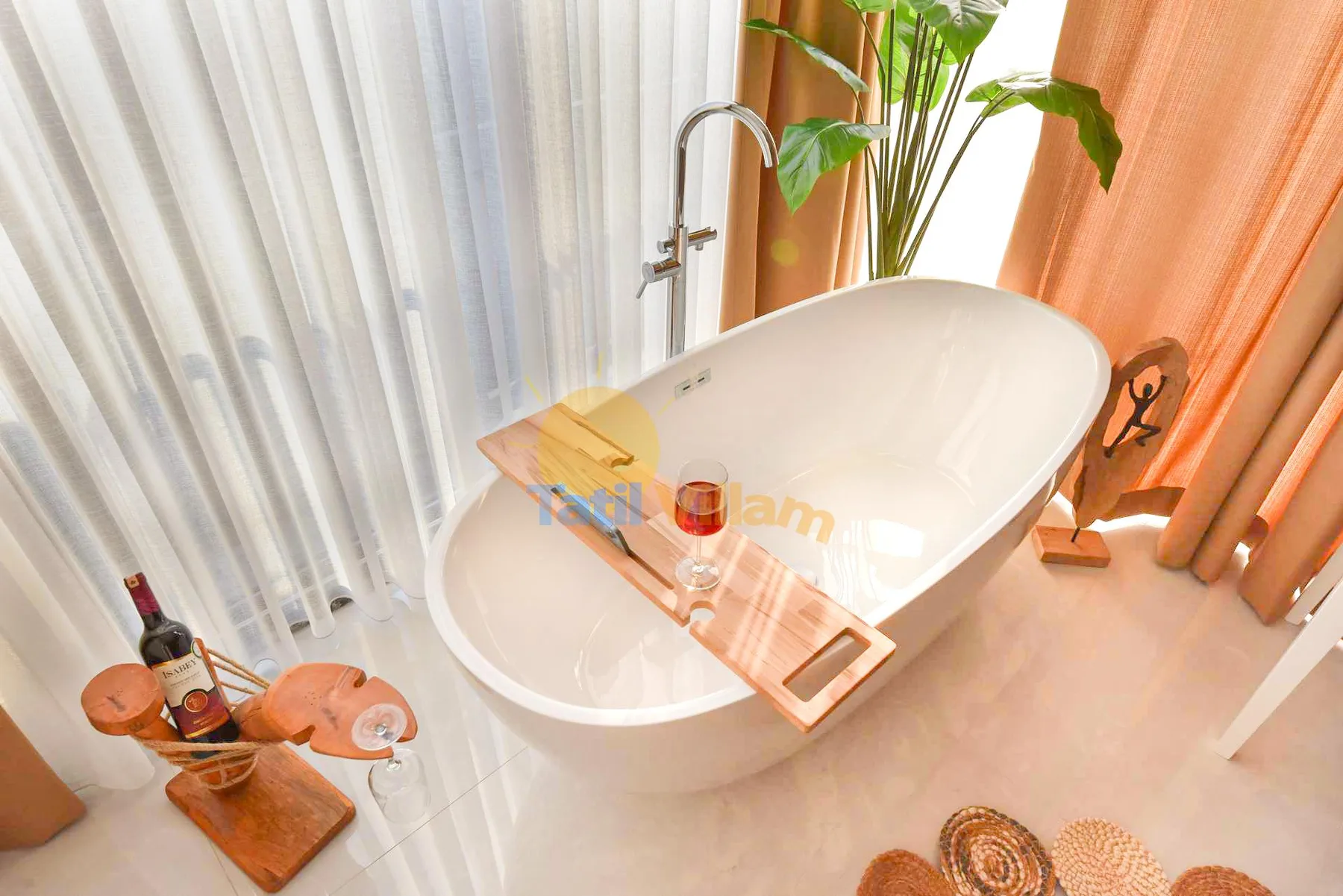 A freestanding white bathtub sits beside sheer curtains and orange drapes, with a leafy plant nearby.