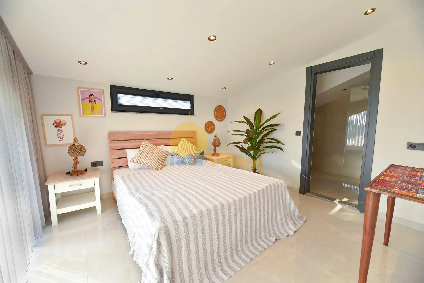 A bright bedroom features a striped bed, white nightstand, framed art, and a potted plant.