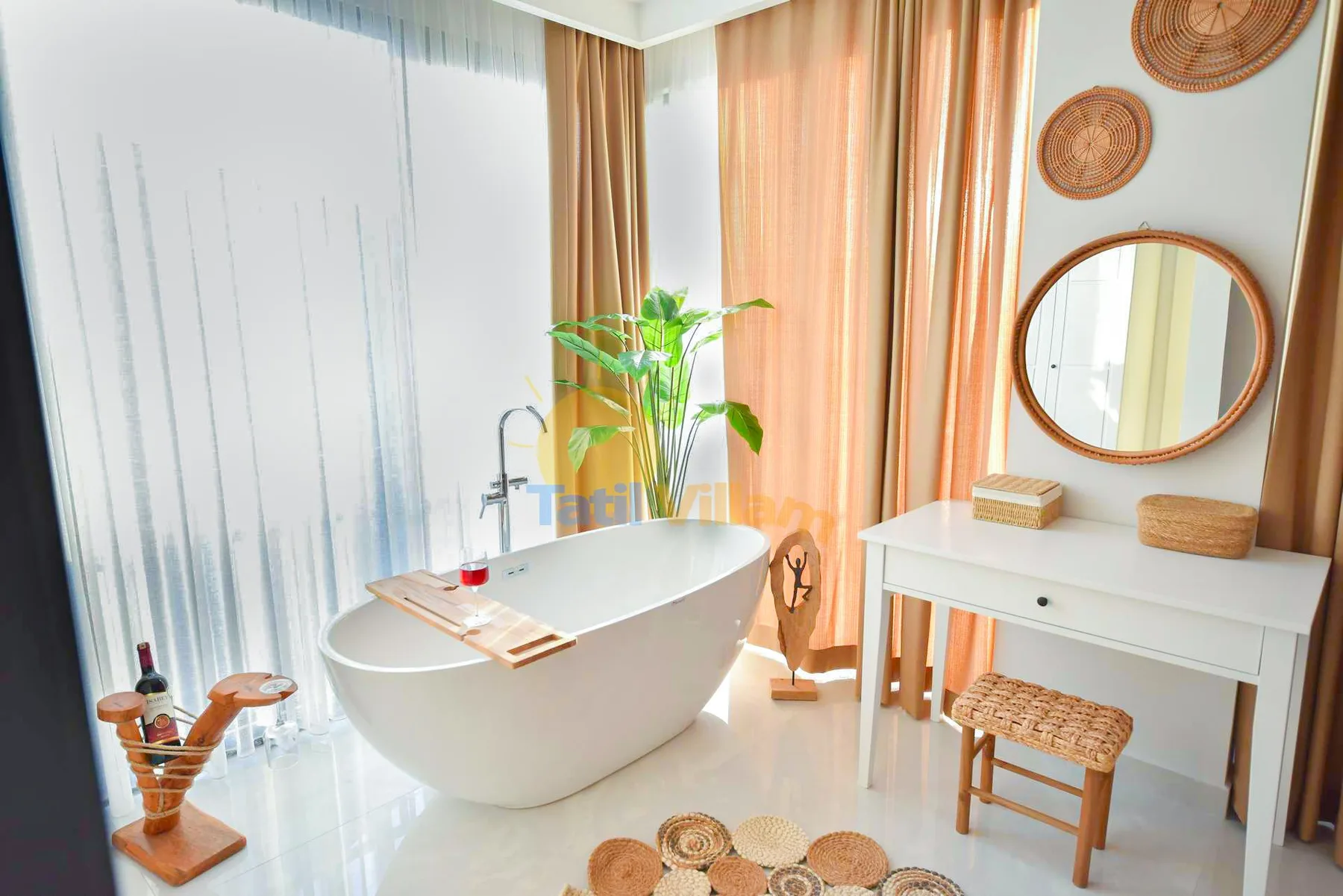 A freestanding white bathtub sits beside sheer curtains, with a white vanity, round mirror, and woven accents.