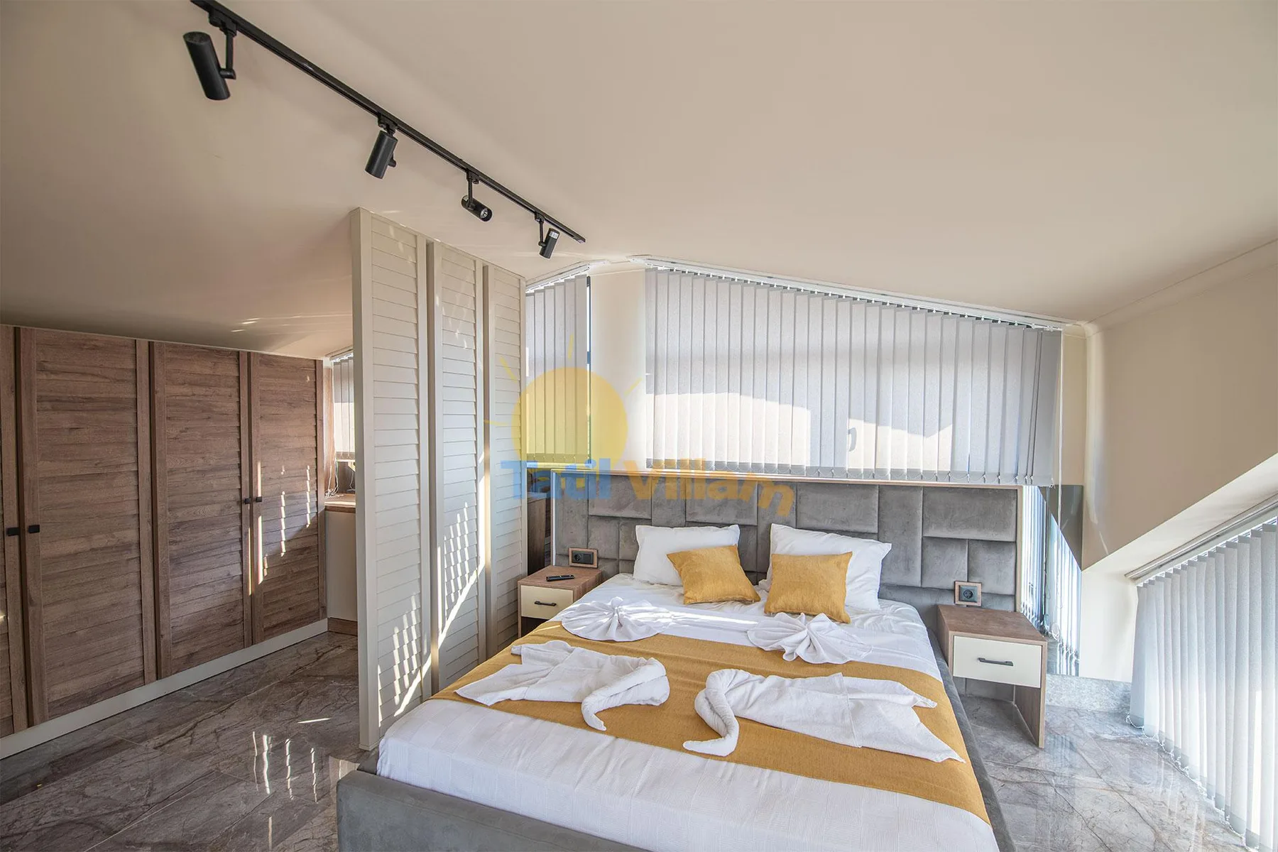 A bedroom with a neatly made bed, mustard-colored pillows, bedside tables, and vertical blinds.