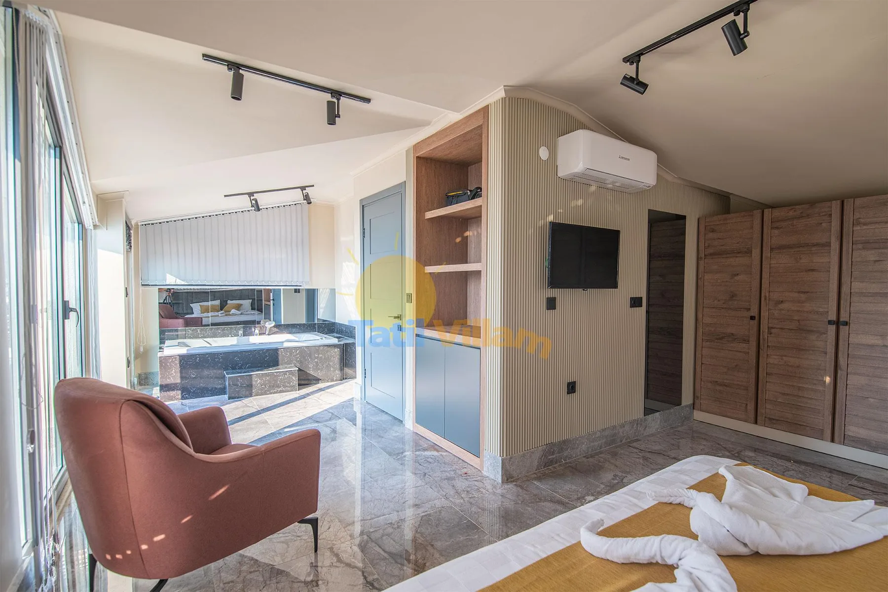 A bright bedroom with a pink armchair, wood wardrobes, and a bed beside floor-to-ceiling windows.