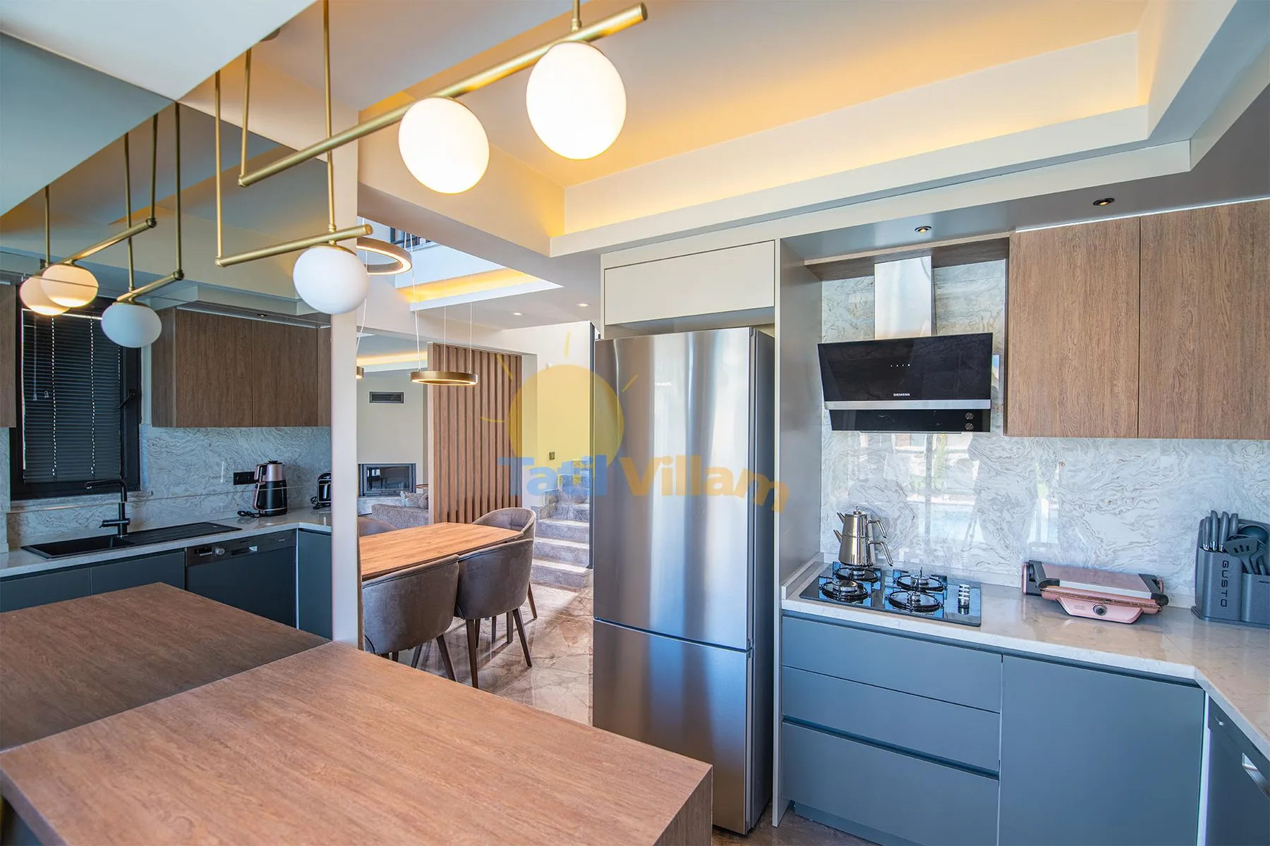 A modern kitchen features wood cabinetry, a stainless-steel refrigerator, a gas cooktop, and a dining table.