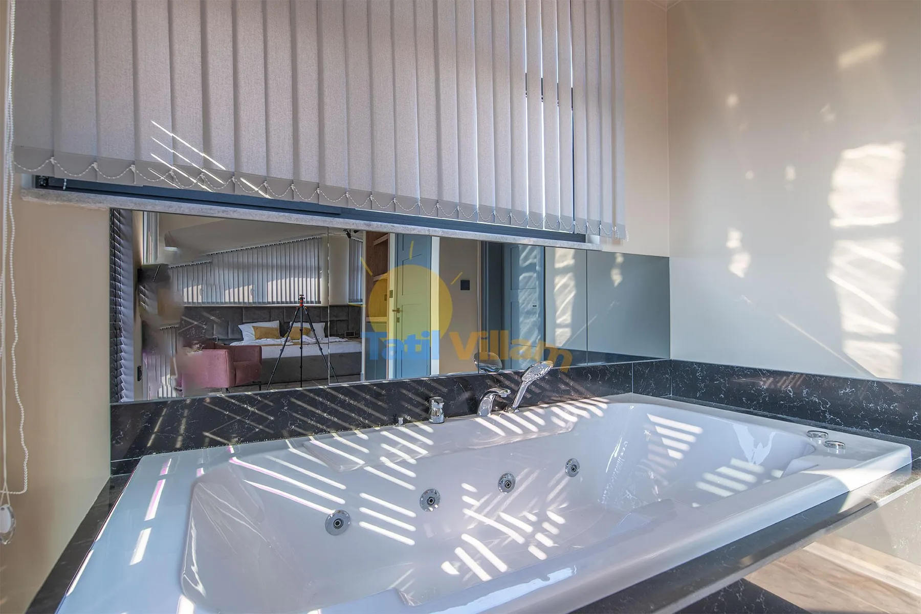A large white whirlpool tub sits beneath a mirrored wall, with sunlight filtering through vertical blinds.