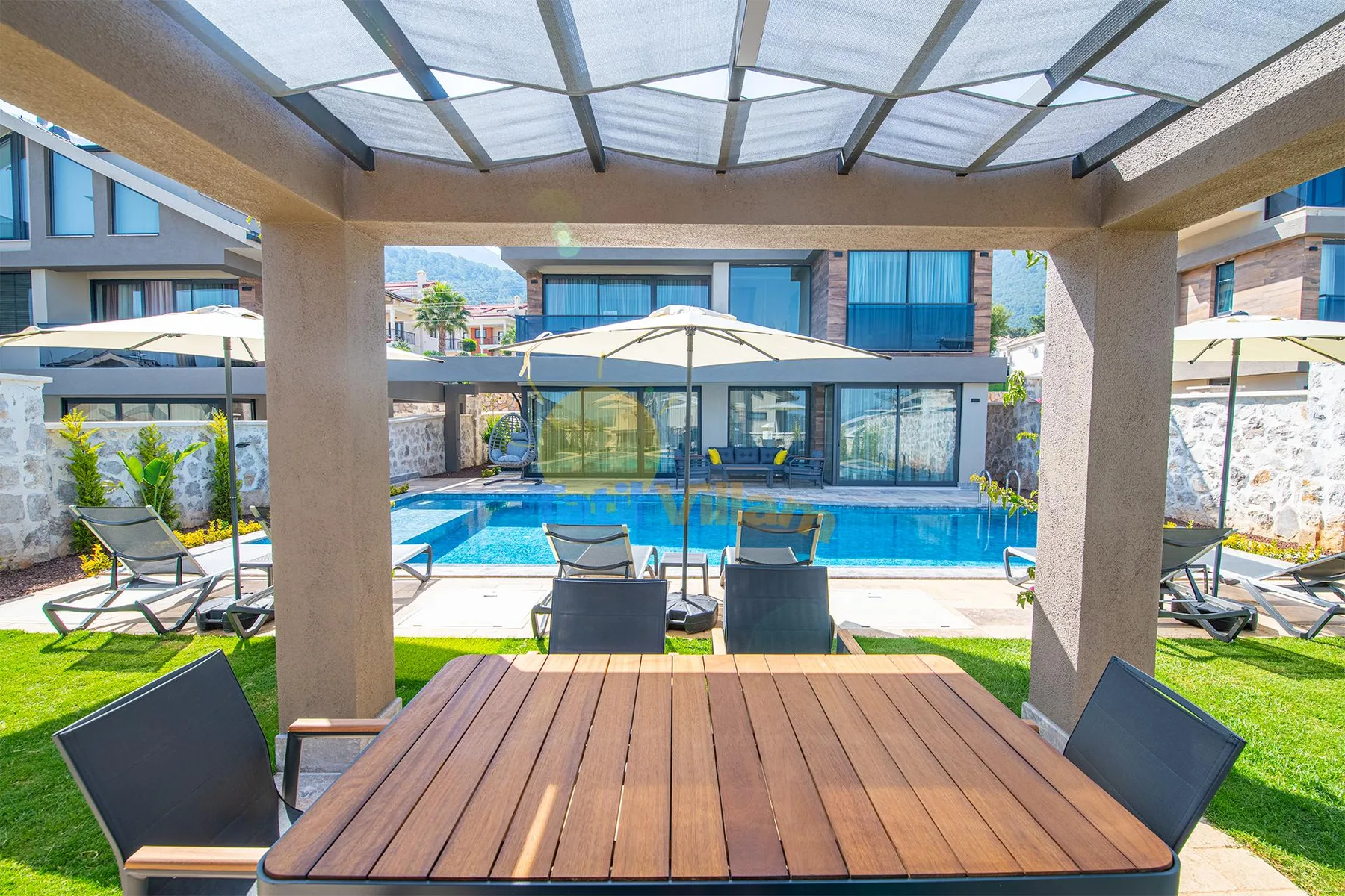 A covered patio with a wooden dining table overlooks a swimming pool and modern glass-fronted building.