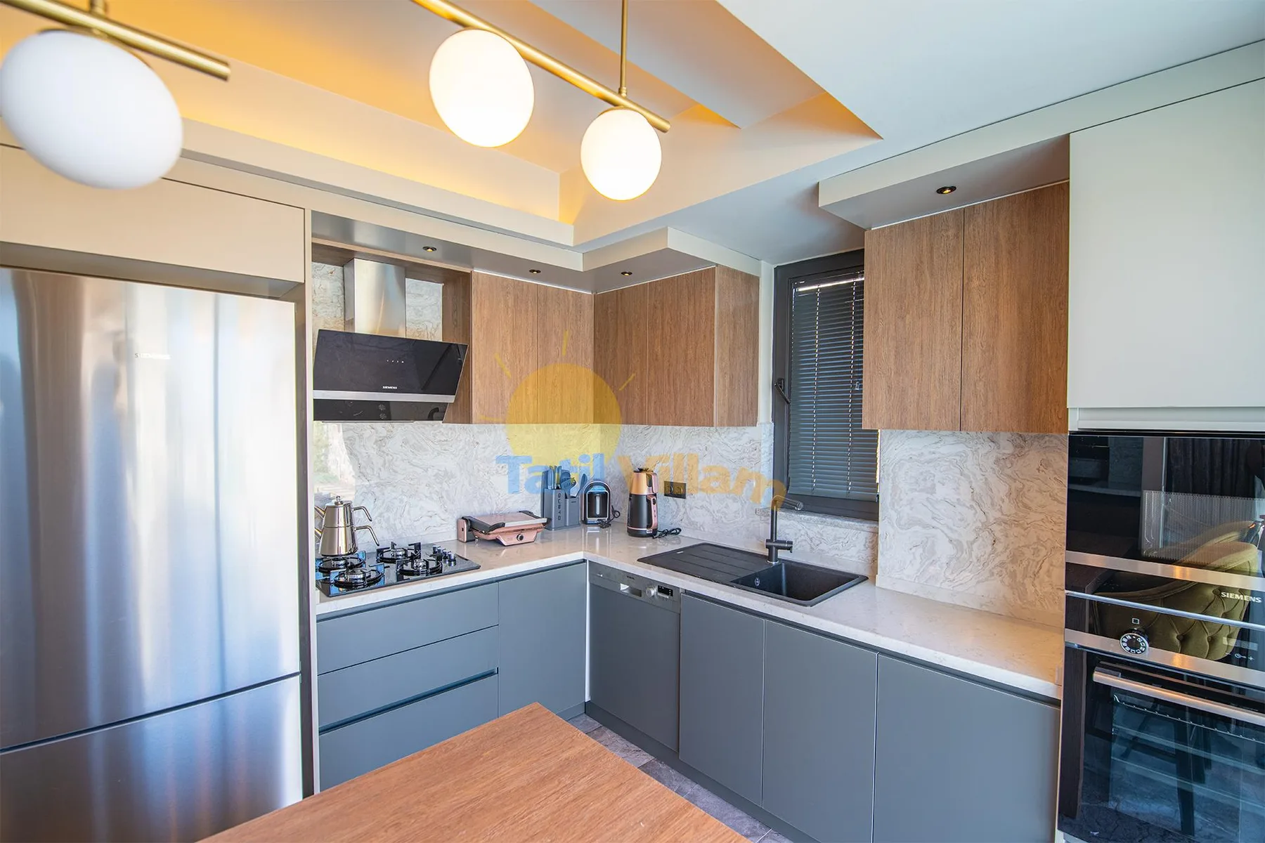 A modern kitchen features stainless steel appliances, wood cabinets, gray counters, and three pendant lights.