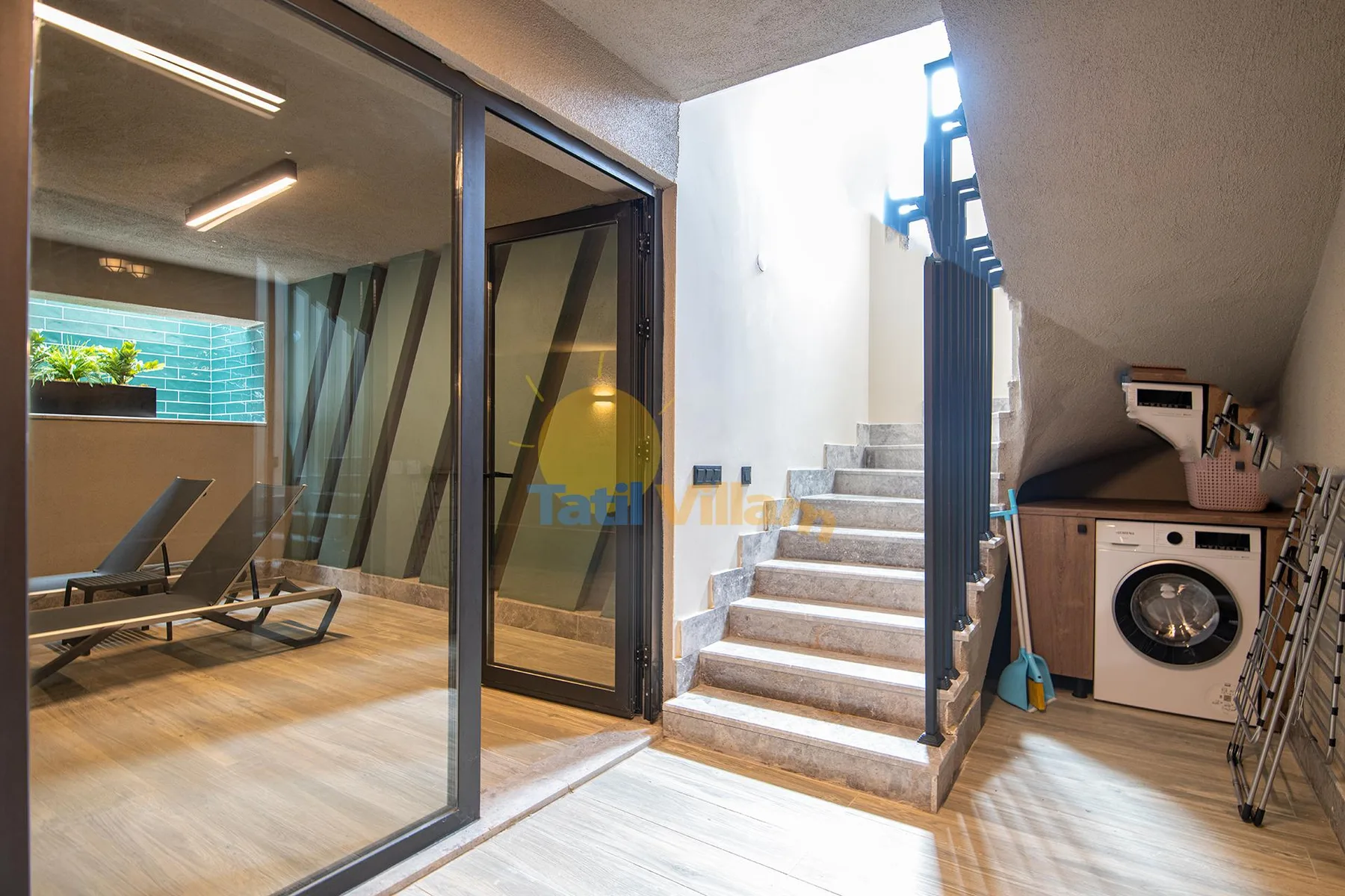 A modern interior shows glass doors beside a staircase, with a washing machine and lounge chairs nearby.