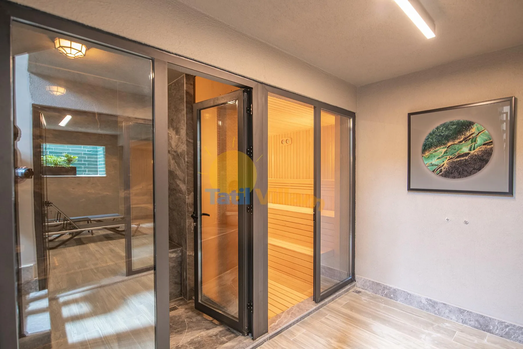 A glass-fronted sauna with warm wood benches sits beside a tiled room and framed wall art.