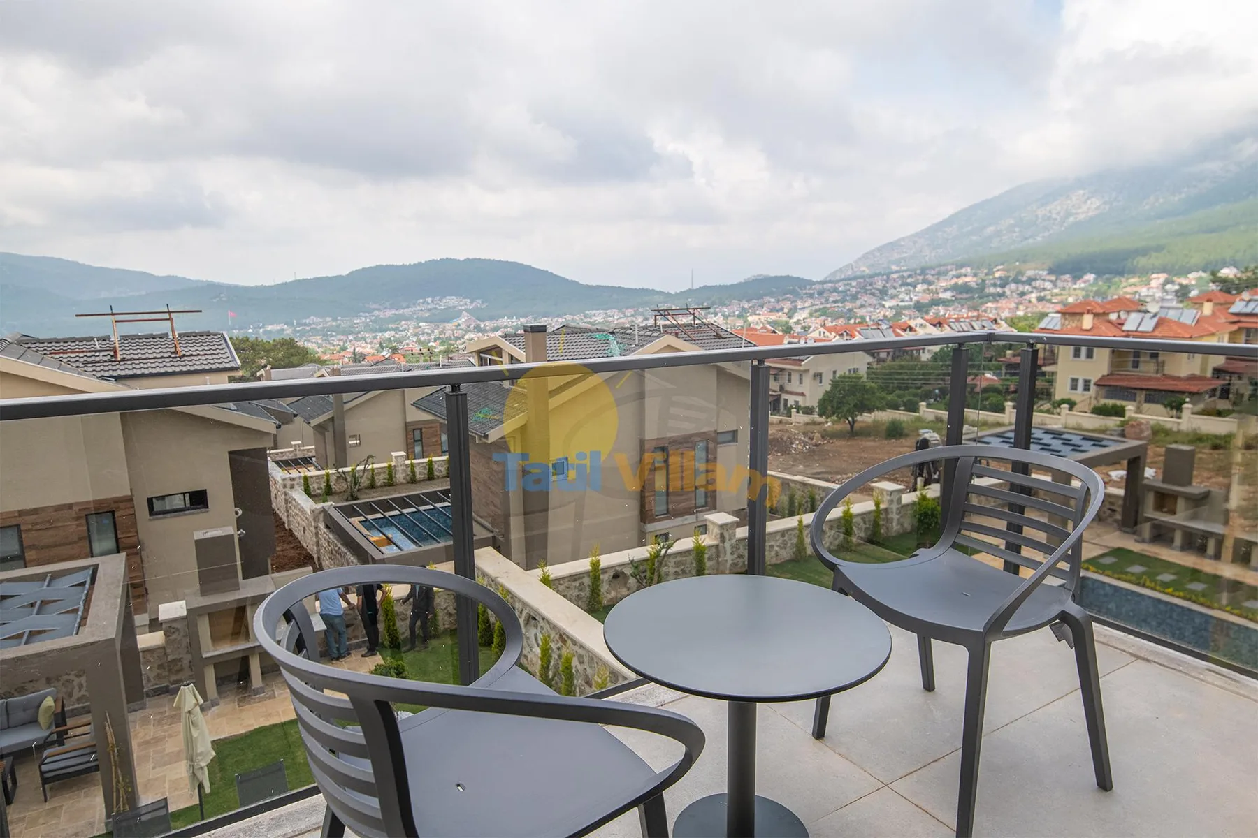 A rooftop terrace has a round table and chairs beside a glass railing, overlooking nearby buildings and mountains.