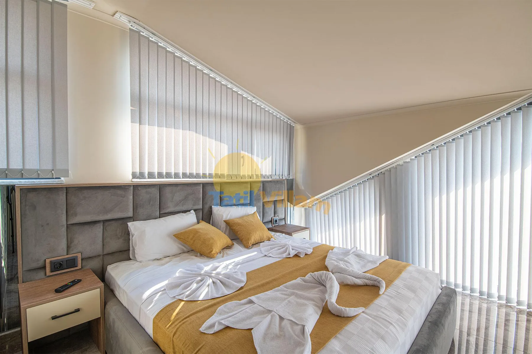 A bedroom with a large bed, mustard-yellow bedding, gray upholstered headboard, and floor-to-ceiling vertical blinds.