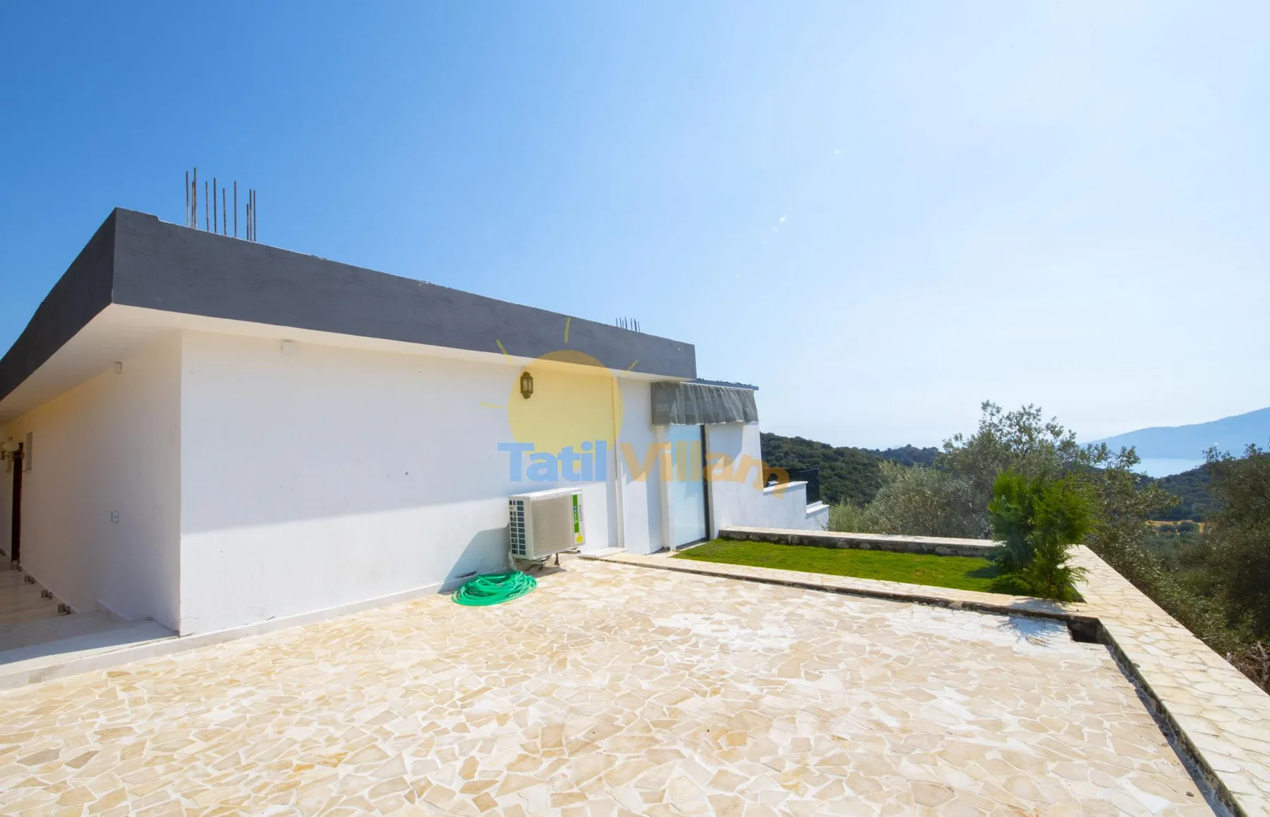A modern house opens onto a broad stone-paved terrace with green hills beyond.
