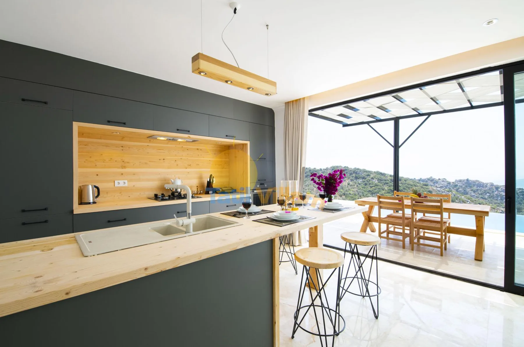 A modern kitchen and dining area open onto a bright terrace with hillside views.