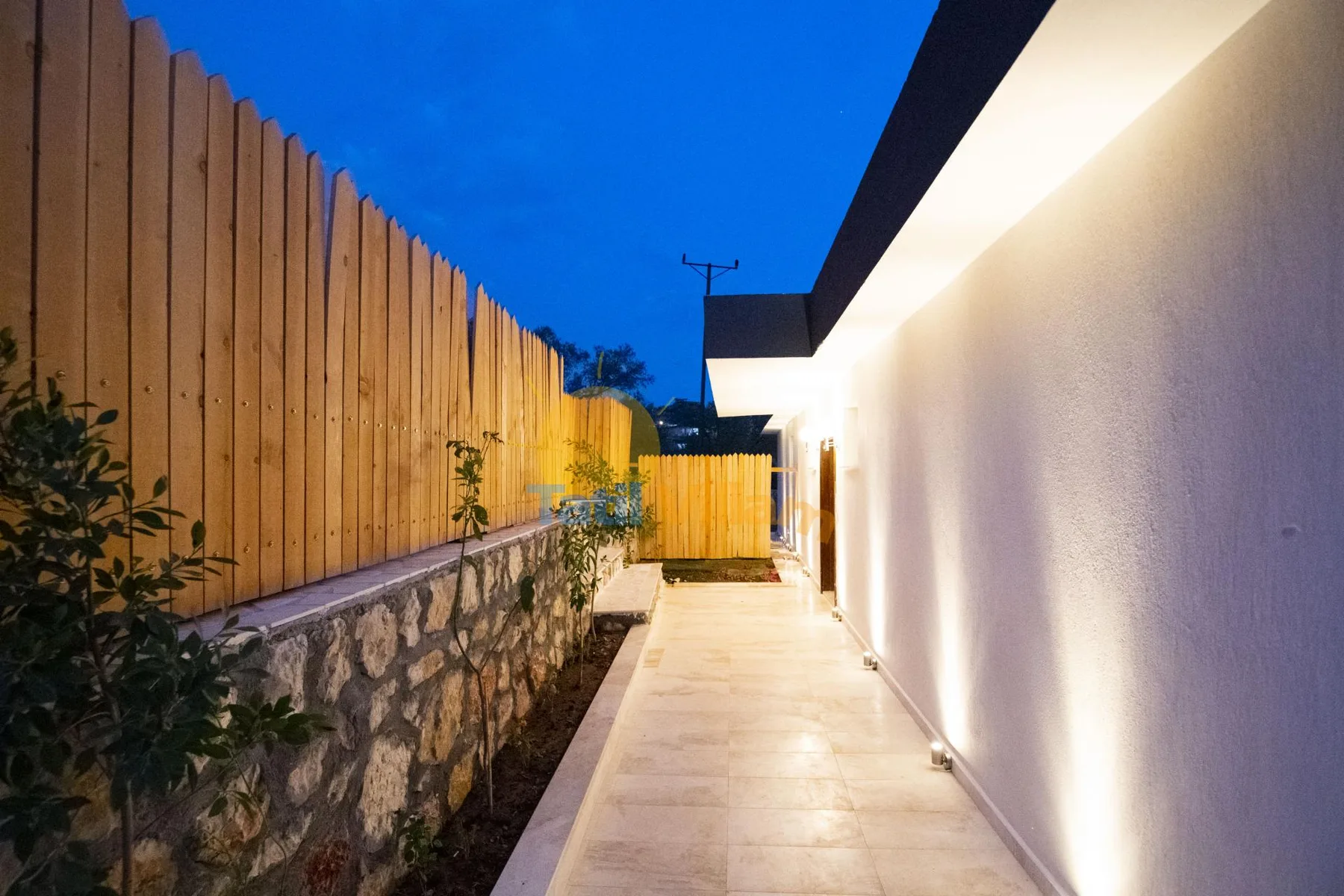 An outdoor walkway runs beside a white building and a wooden fence.
