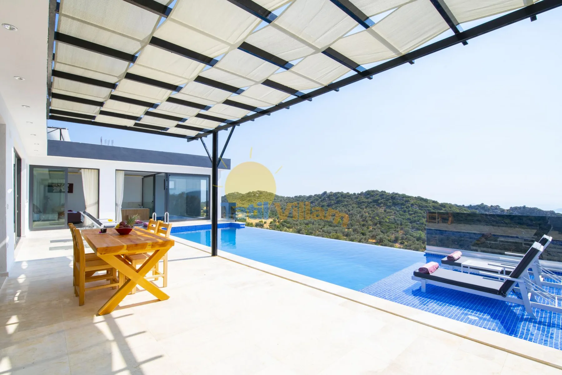 A covered patio with a dining table overlooks a swimming pool and wooded hills.