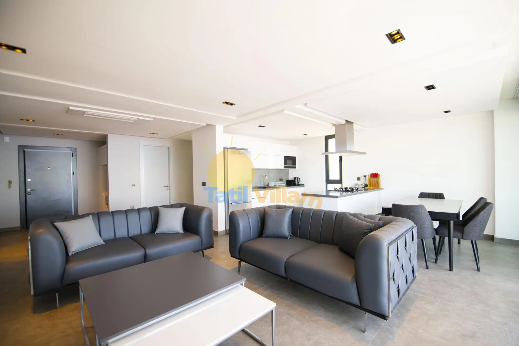 A modern living room with dark seating and an open kitchen in the background.