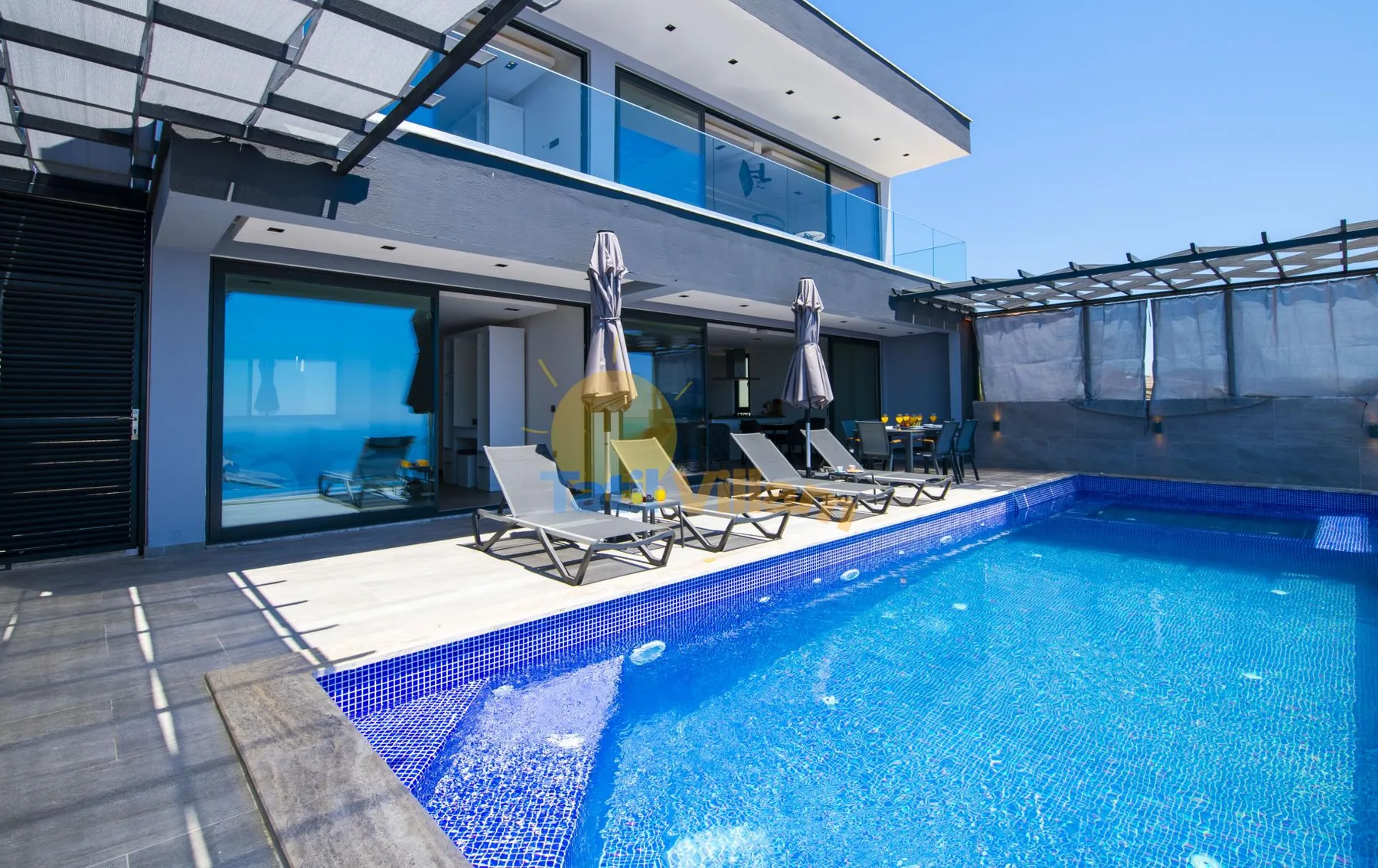 A modern house opens onto a swimming pool with several loungers along the edge.