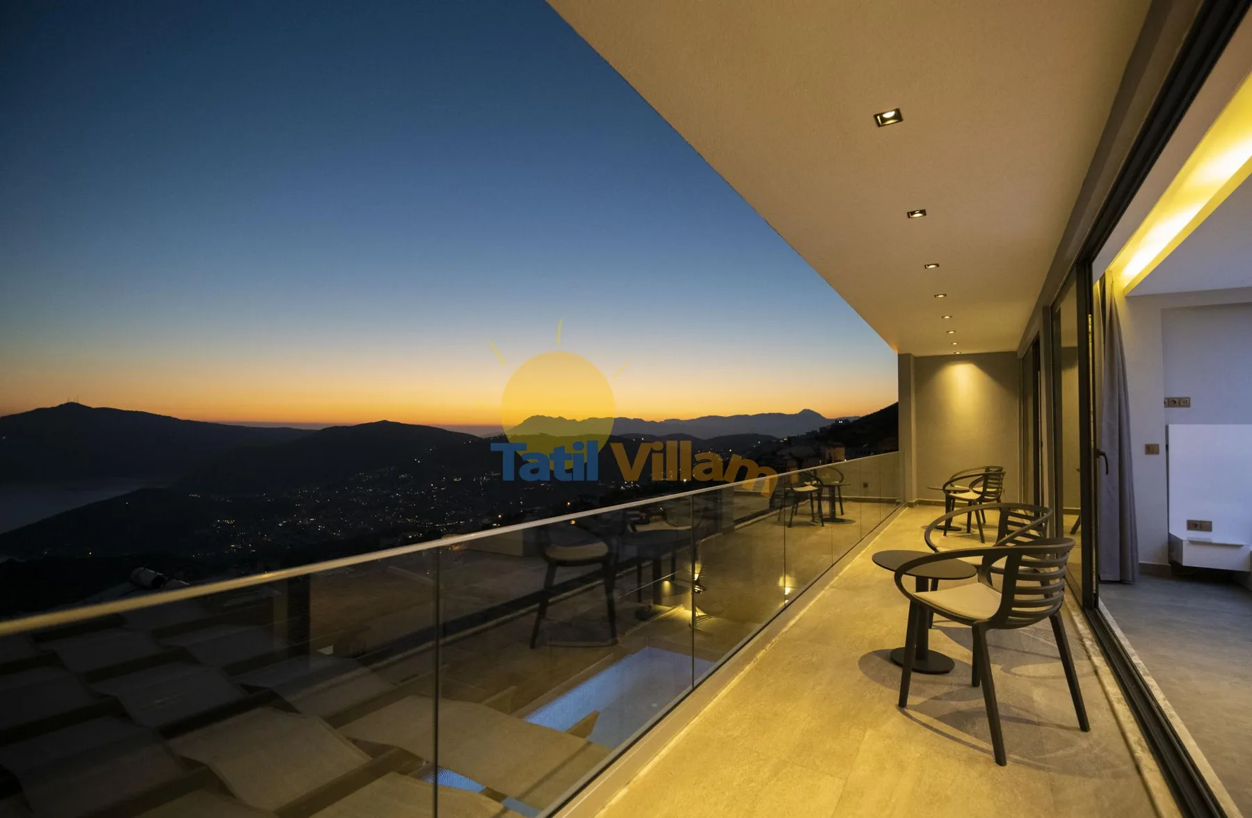 A terrace with seating overlooks mountain silhouettes beneath a colorful sunset sky.
