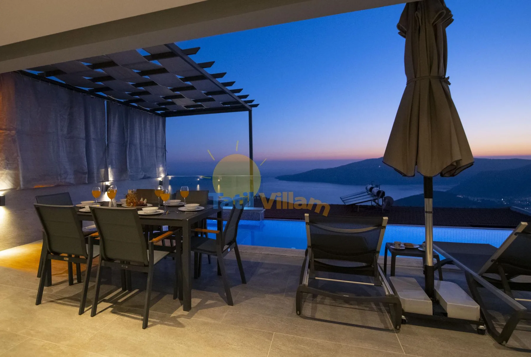A covered outdoor dining area overlooks a pool and the sea at dusk.