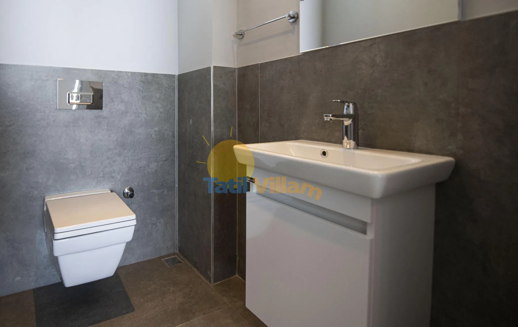 A bathroom with a wall-mounted toilet and a white sink against dark tiled walls.