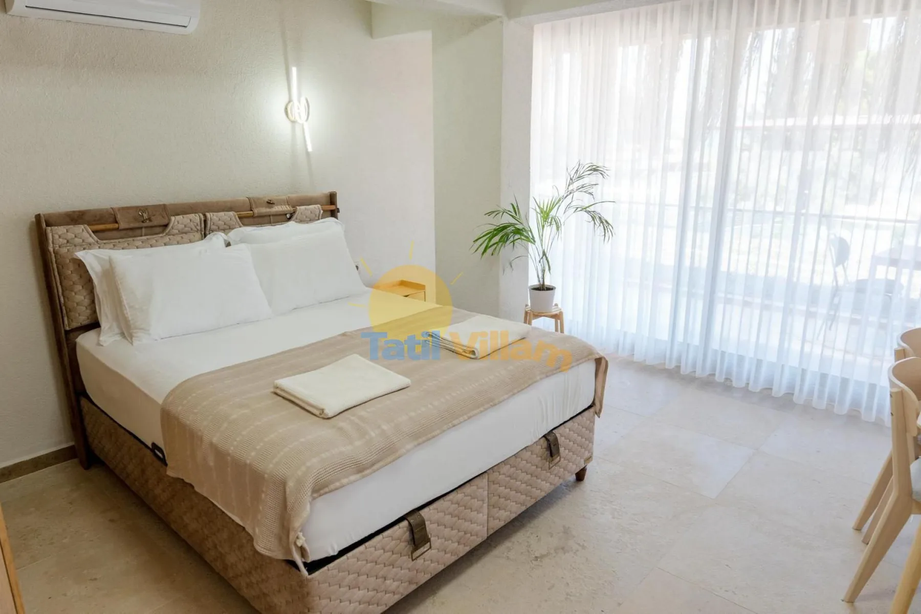 Un dormitorio luminoso con una cama bien tendida junto a una gran ventana con cortinas.
