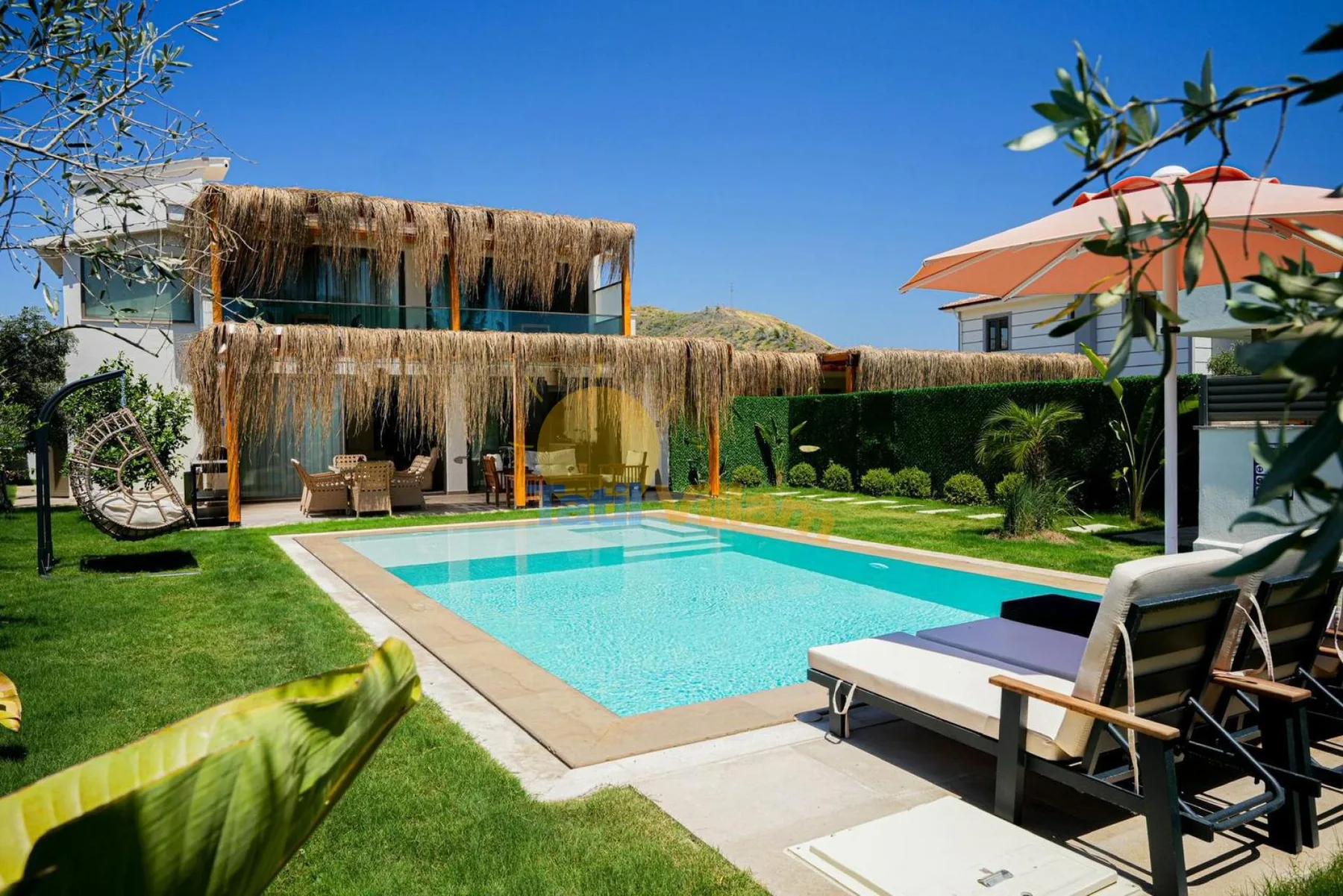 Una piscina al aire libre junto a un césped, asientos y un edificio con techo de paja.