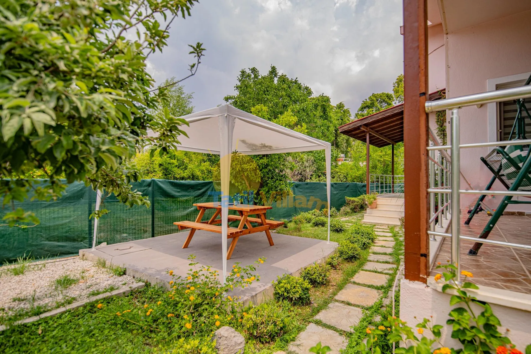 Un patio al aire libre tiene una mesa de picnic bajo una cubierta, junto a un sendero de jardín.