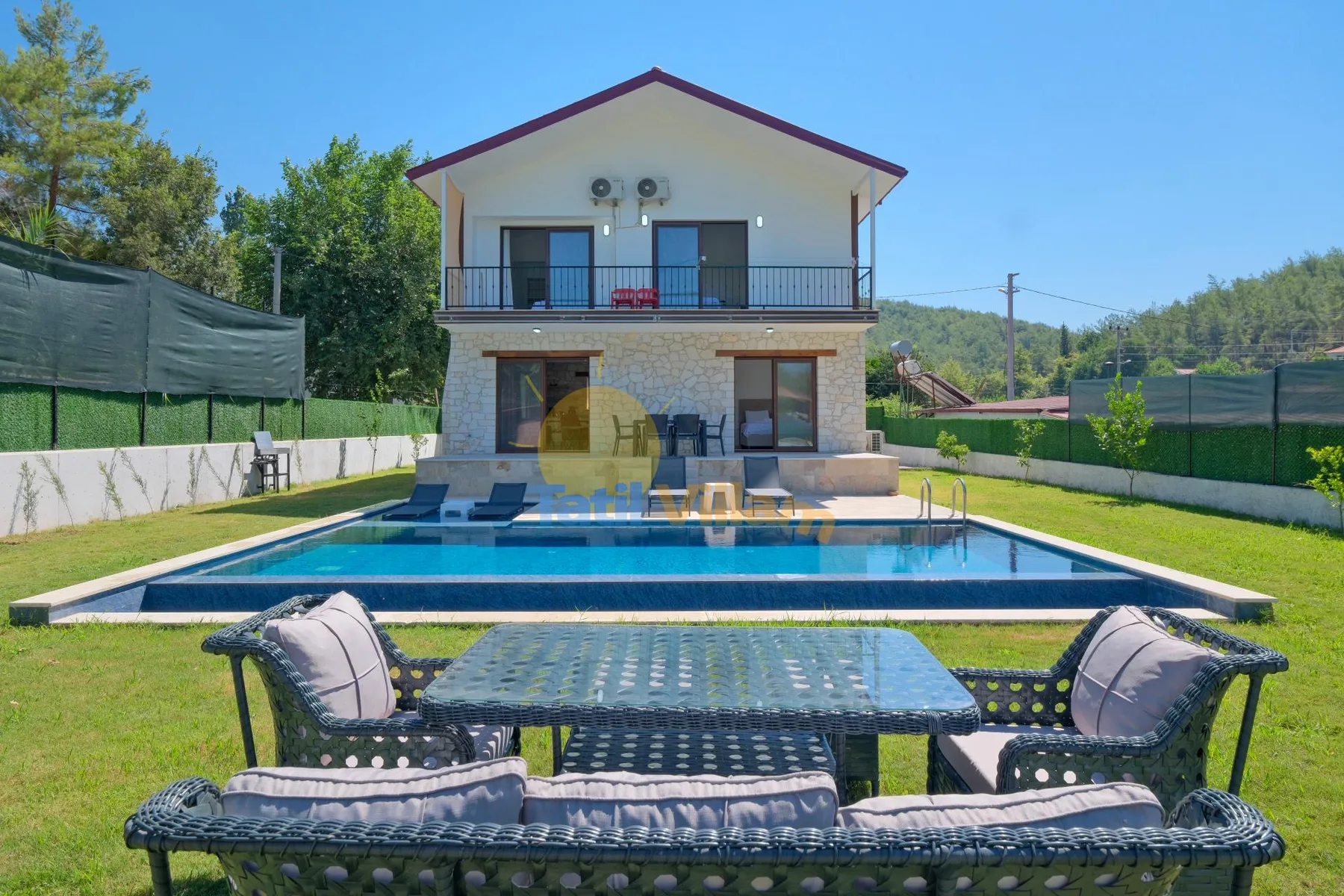 Una casa de dos plantas está detrás de una piscina, con asientos al aire libre en primer plano.