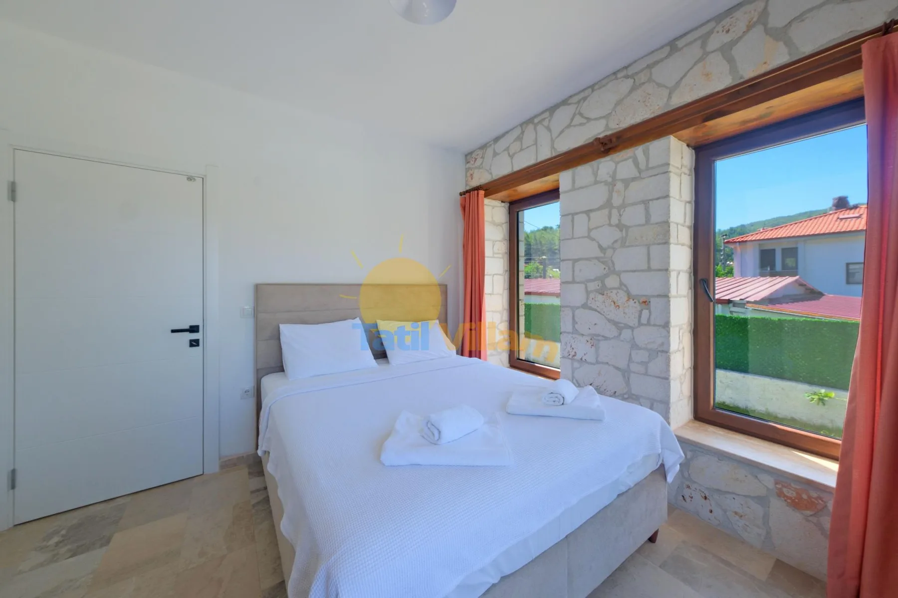 Un dormitorio con una cama bien tendida junto a grandes ventanas y paredes de piedra.