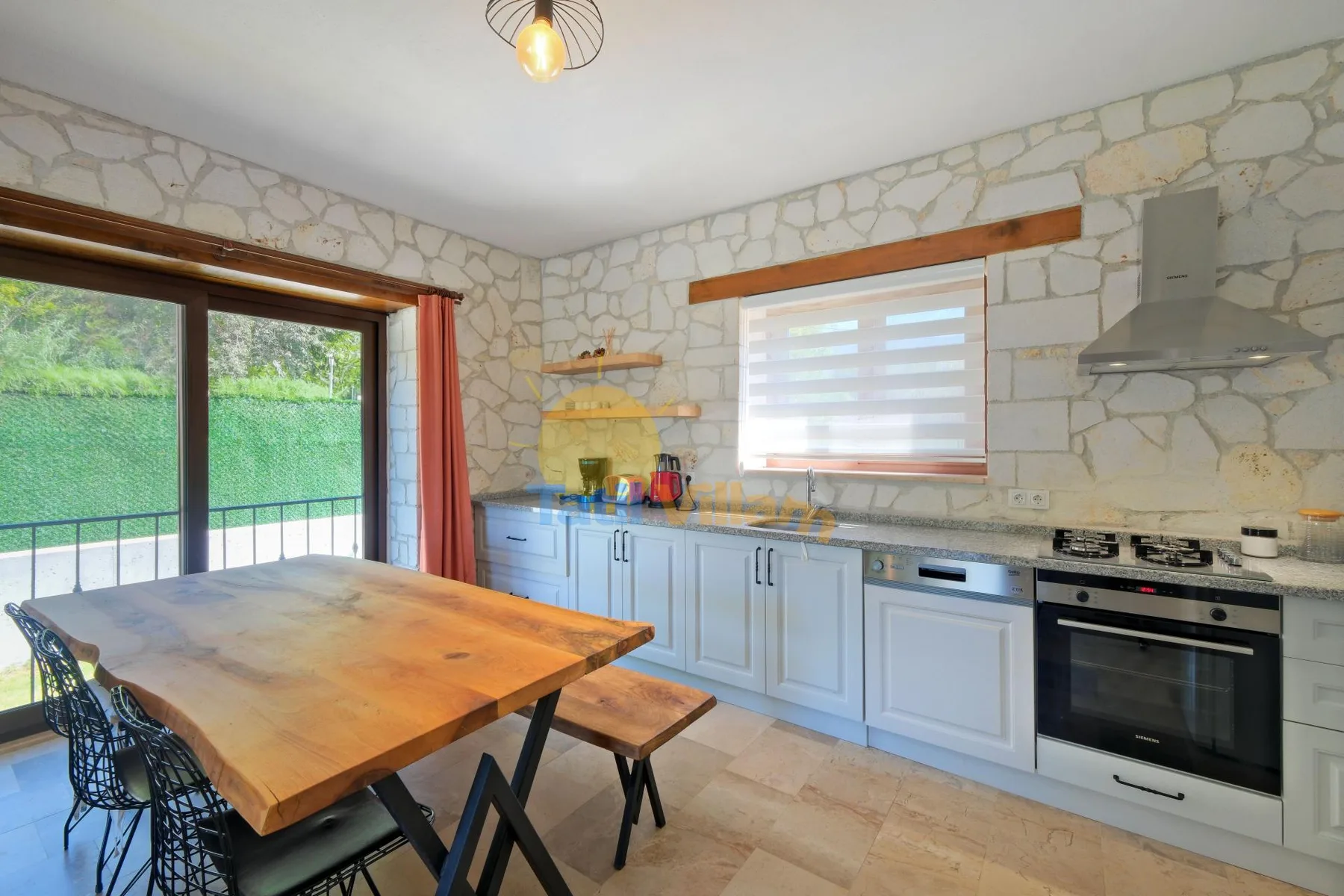 Cocina con paredes de piedra, armarios claros, una mesa de comedor y ventanas grandes.