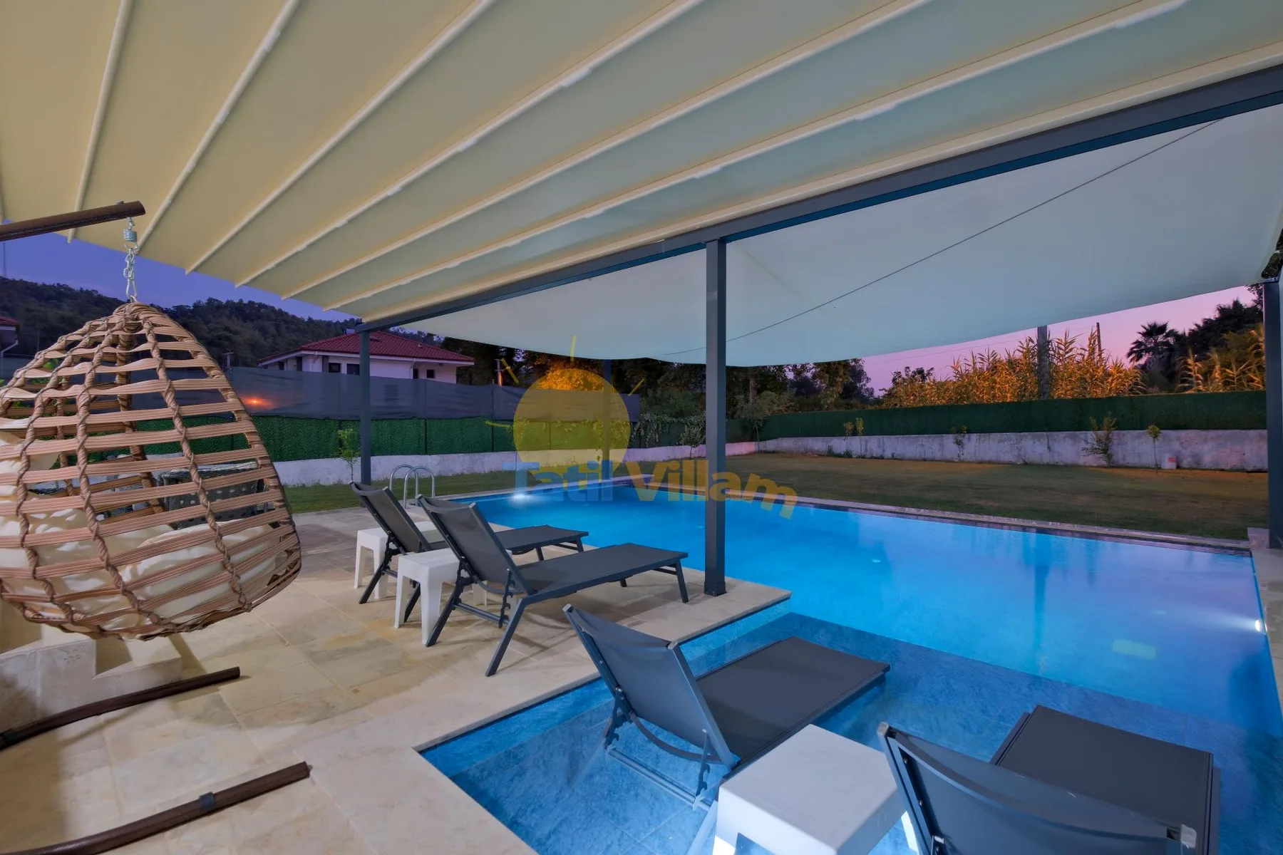 Un patio cubierto con asientos da a una piscina iluminada al anochecer.