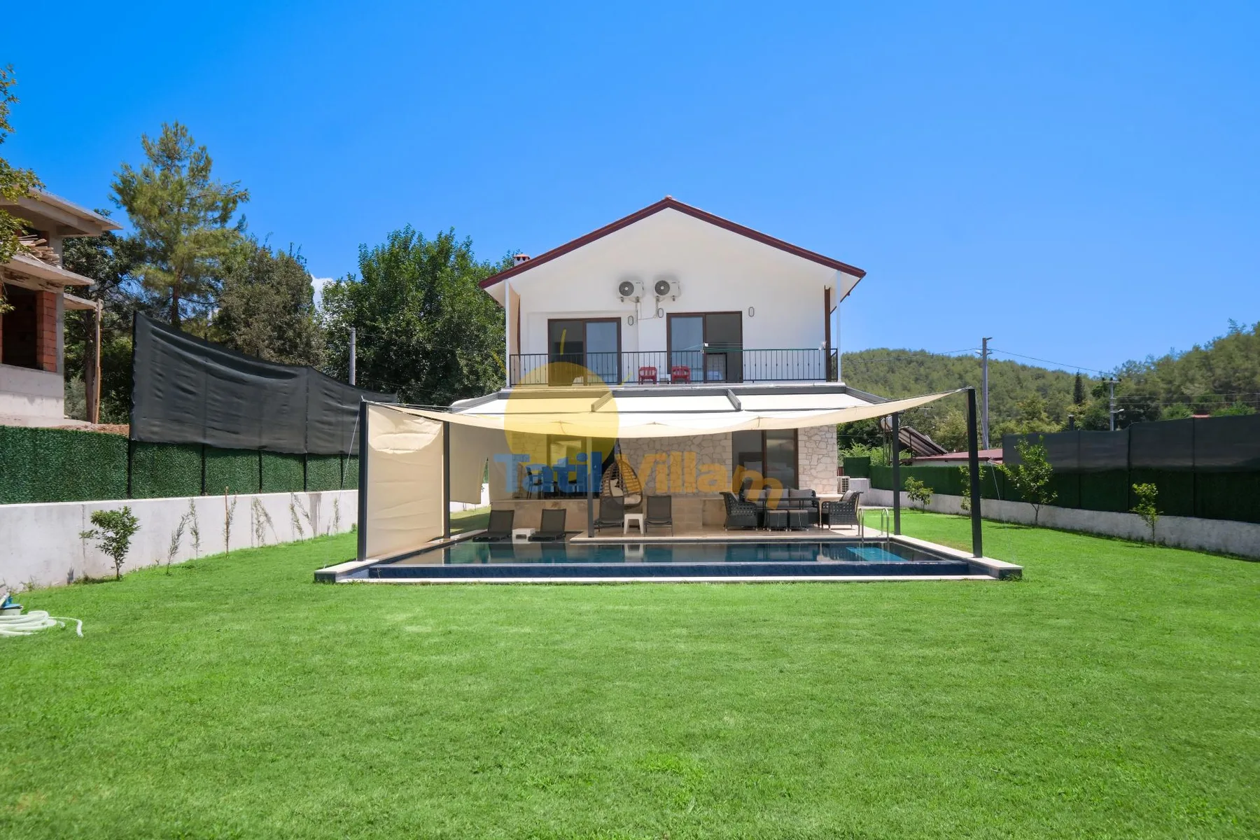 Una casa de dos plantas se alza tras un césped, con patio sombreado y piscina.