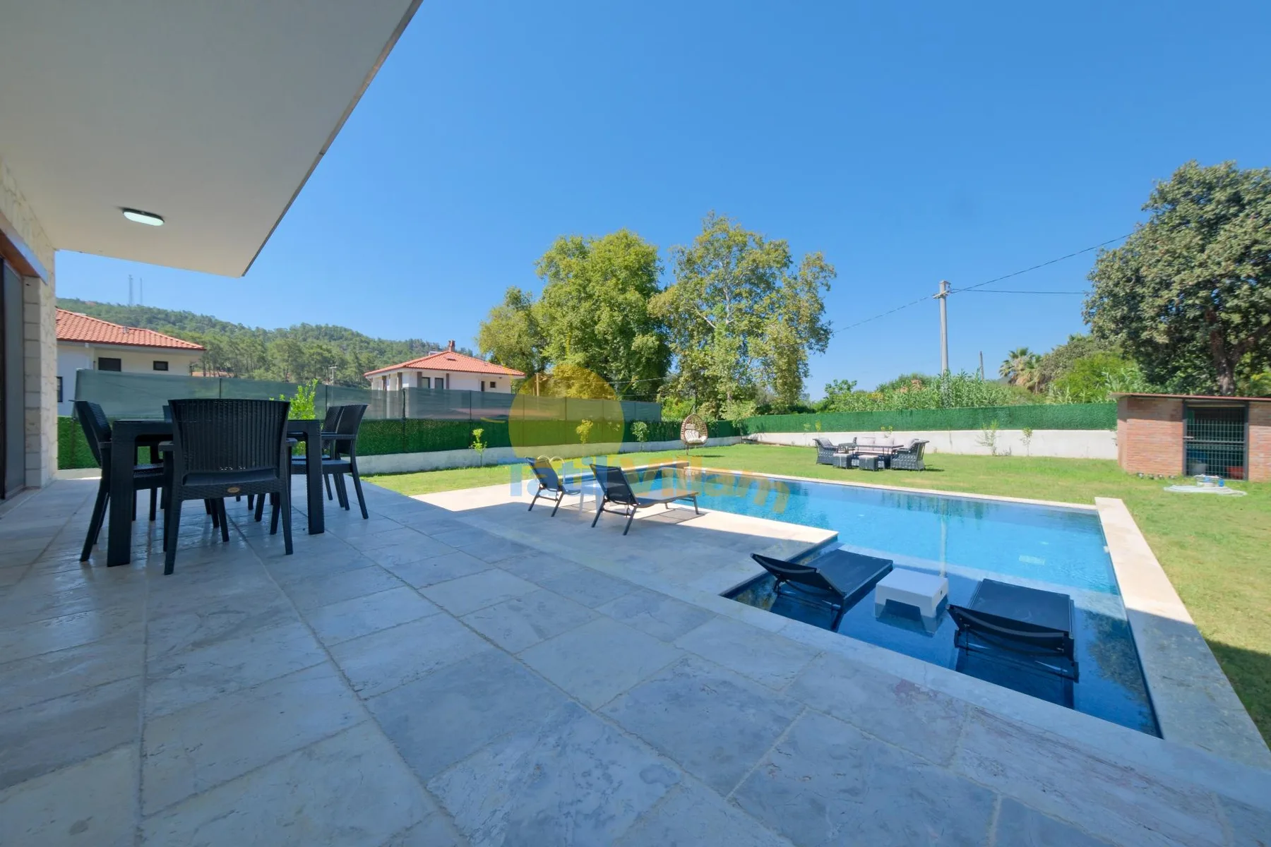Un patio cubierto da a una piscina, un césped y asientos al aire libre.